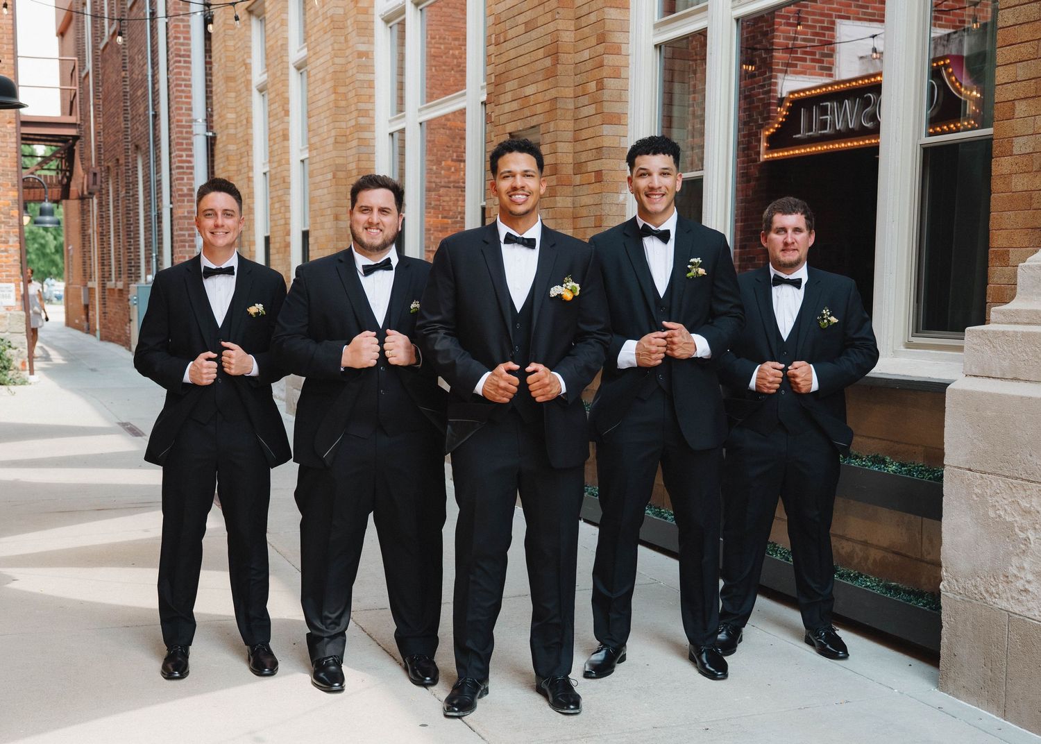 Five groomsmen in black tuxedos pose together on steps outside a brick building.