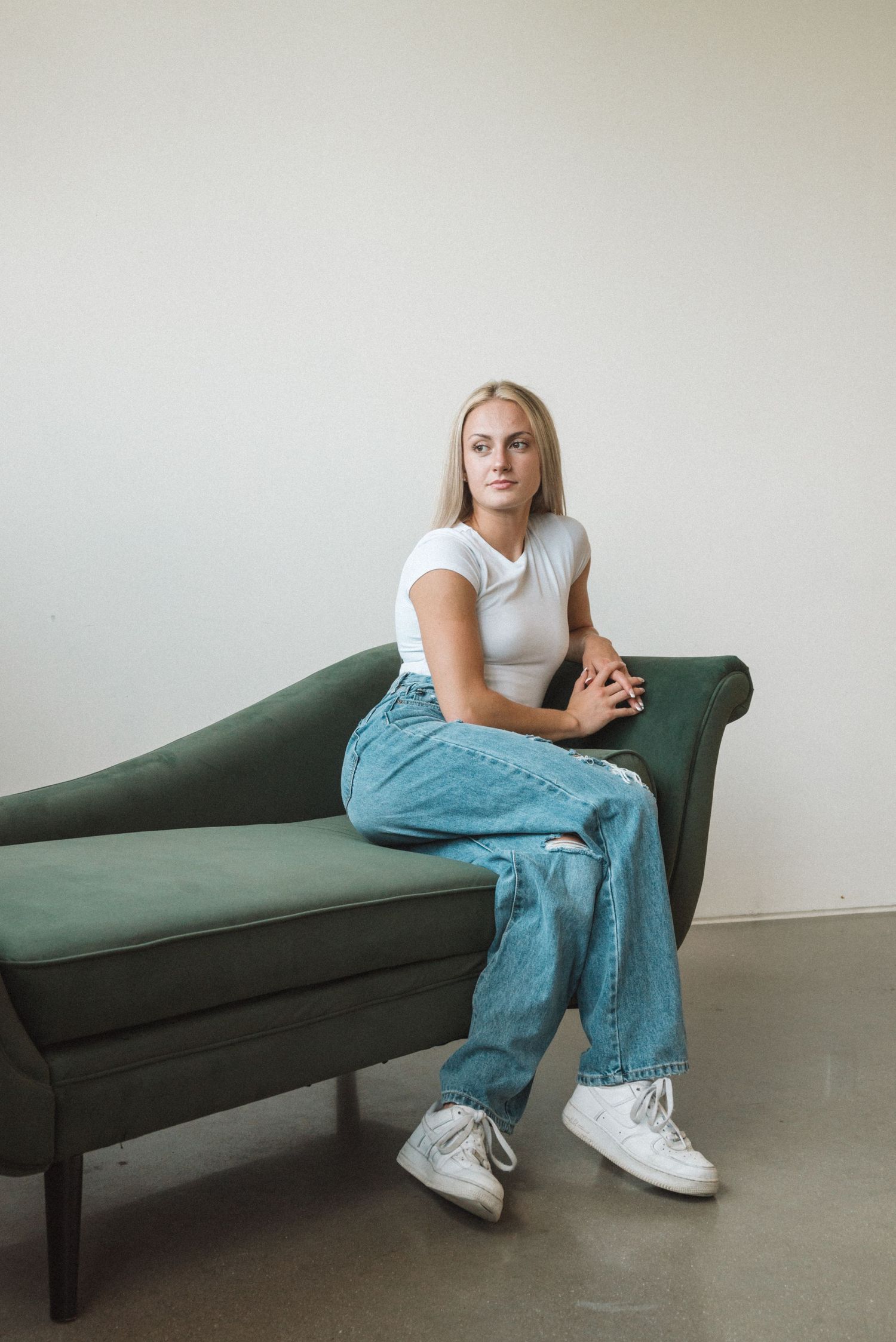 Person in casual outfit sitting on dark green vintage couch against white wall.