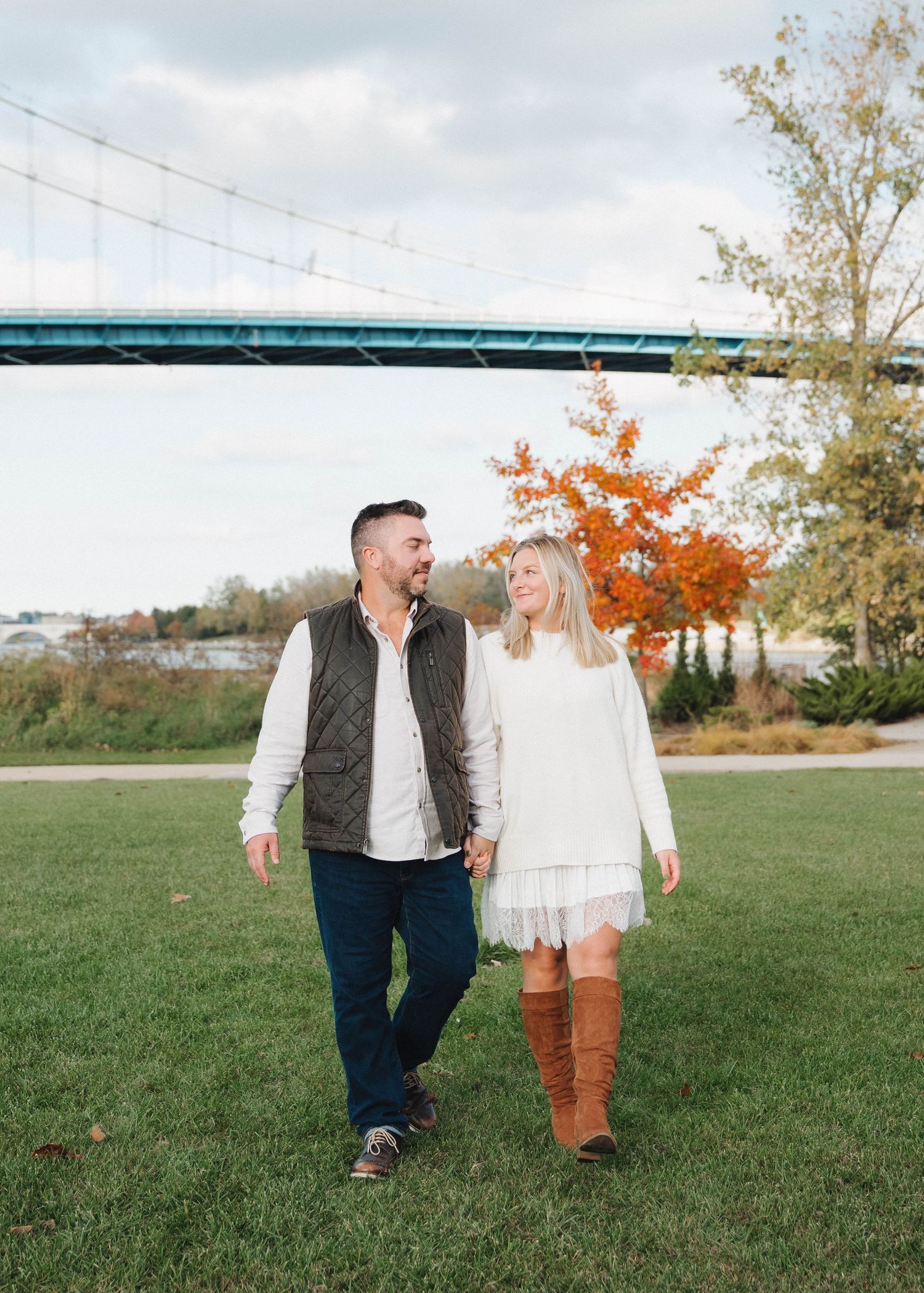 Couple walking together on grassy field during autumn with orange foliage in the background.