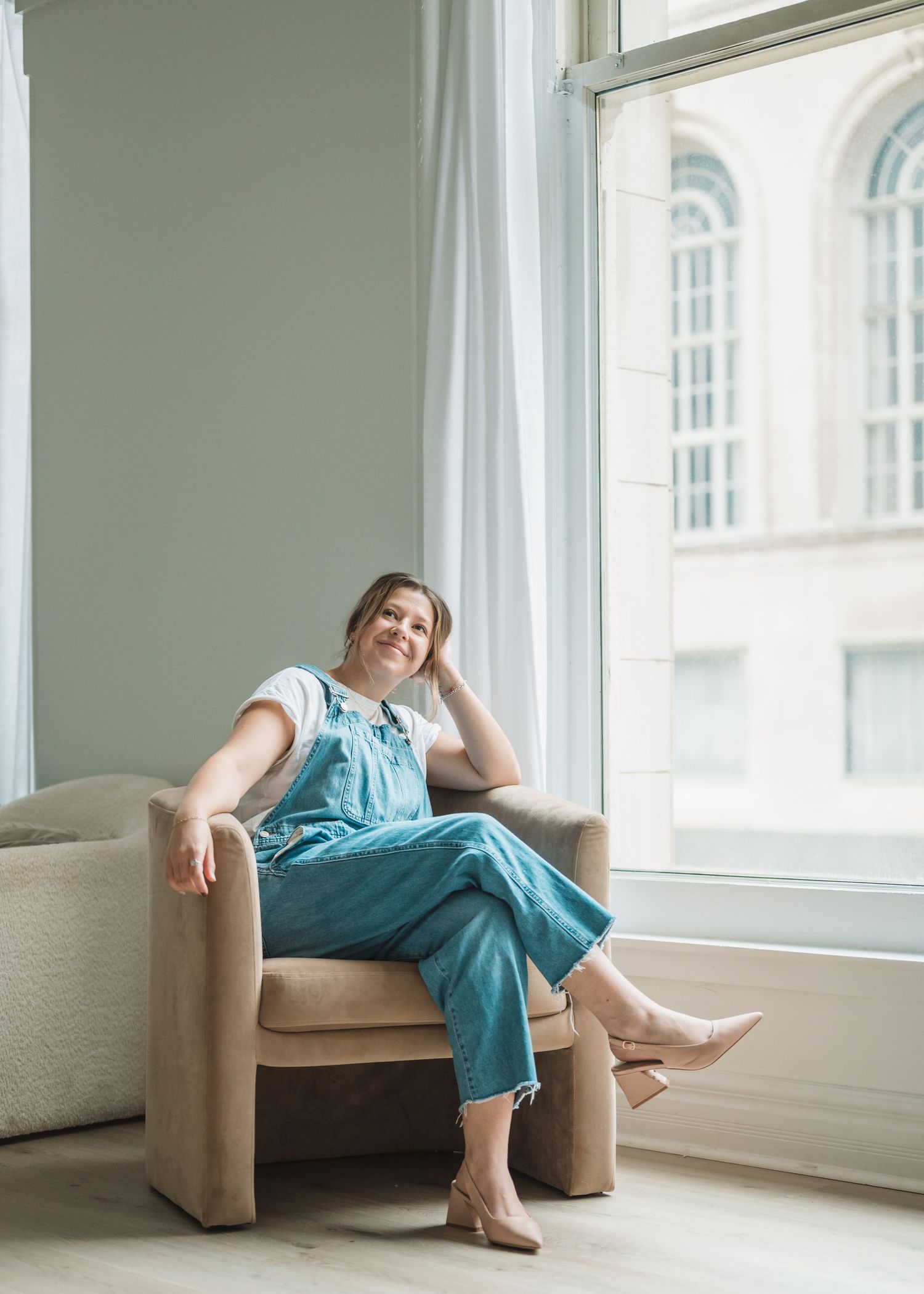 Person lounging in blue overalls on beige armchair by large window.
