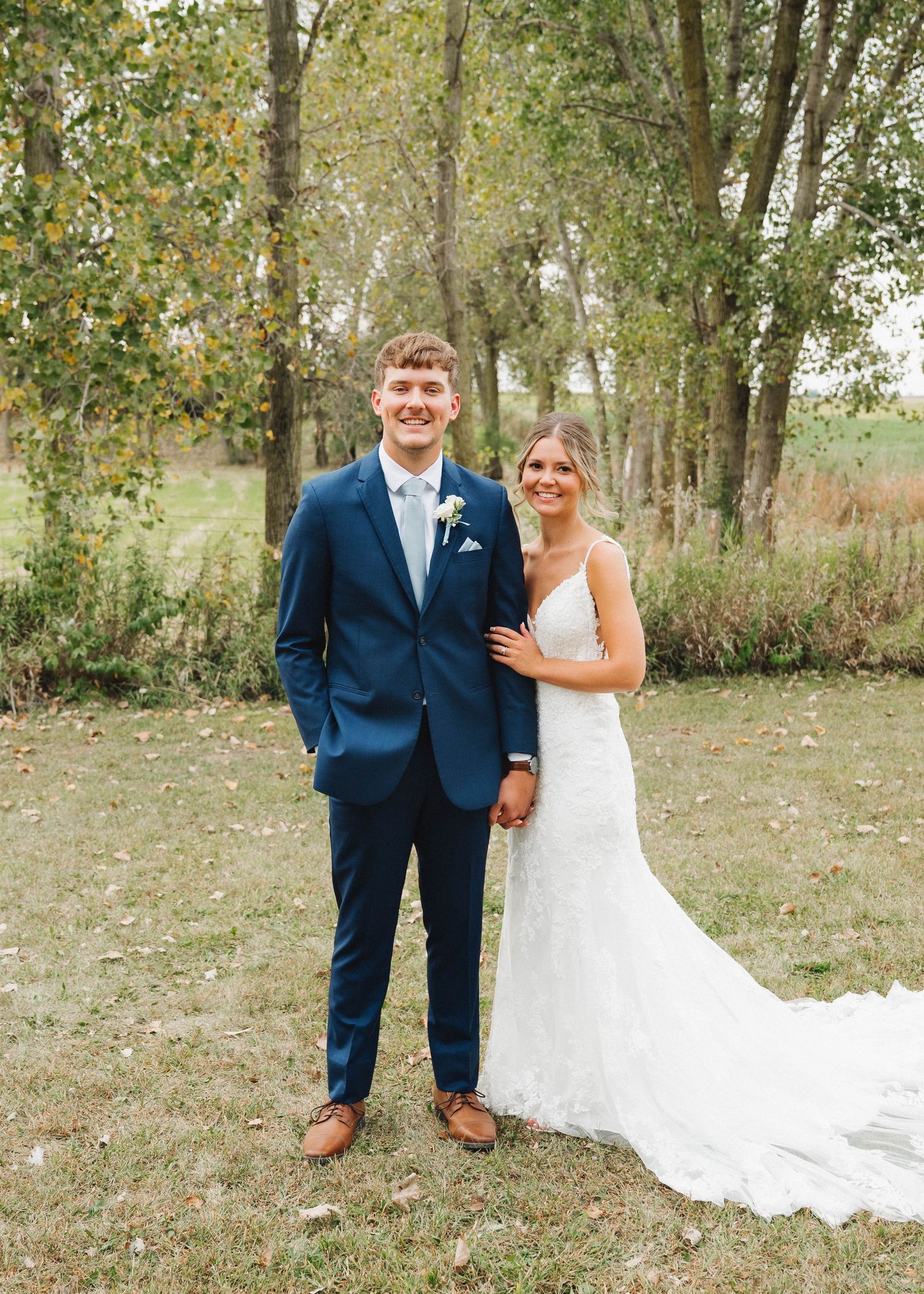 A couple in wedding attire poses for photos in a wooded outdoor setting with autumn trees in the background.