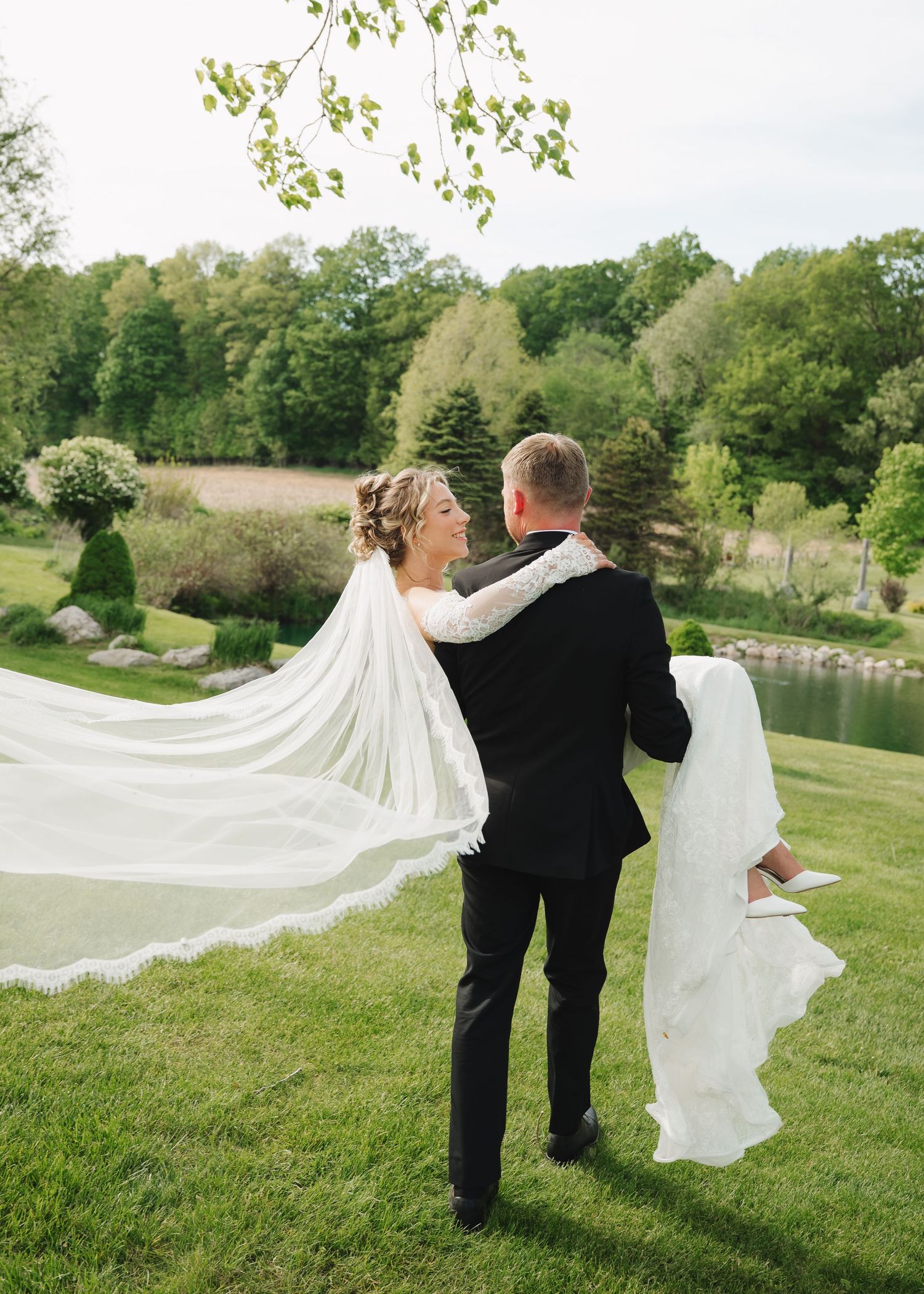 Bride and groom stand together outdoors, her long white veil flowing in the breeze, with lush green trees and a pond behind them.