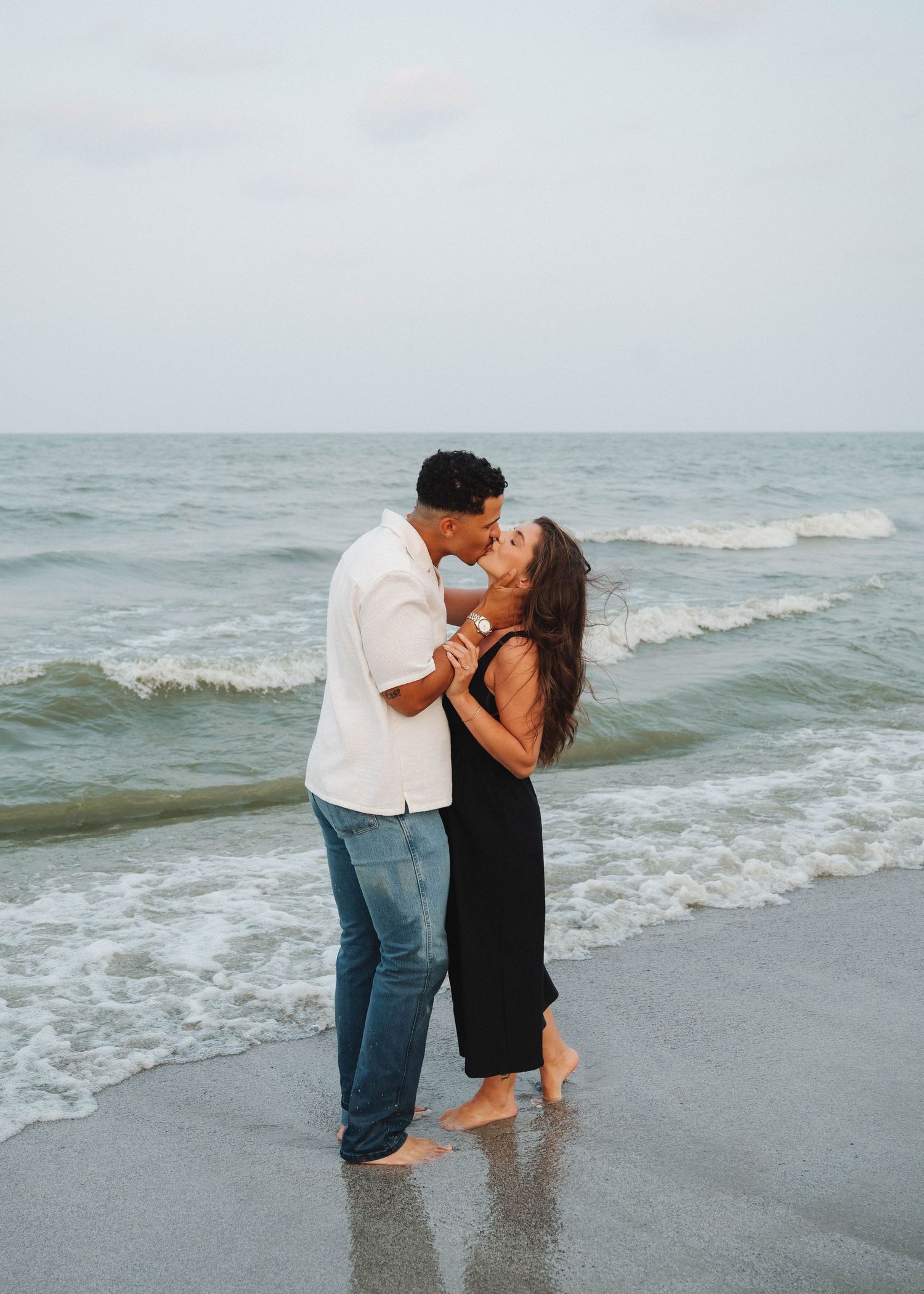 A couple shares a romantic moment while standing in the ocean waves at the beach during sunset.