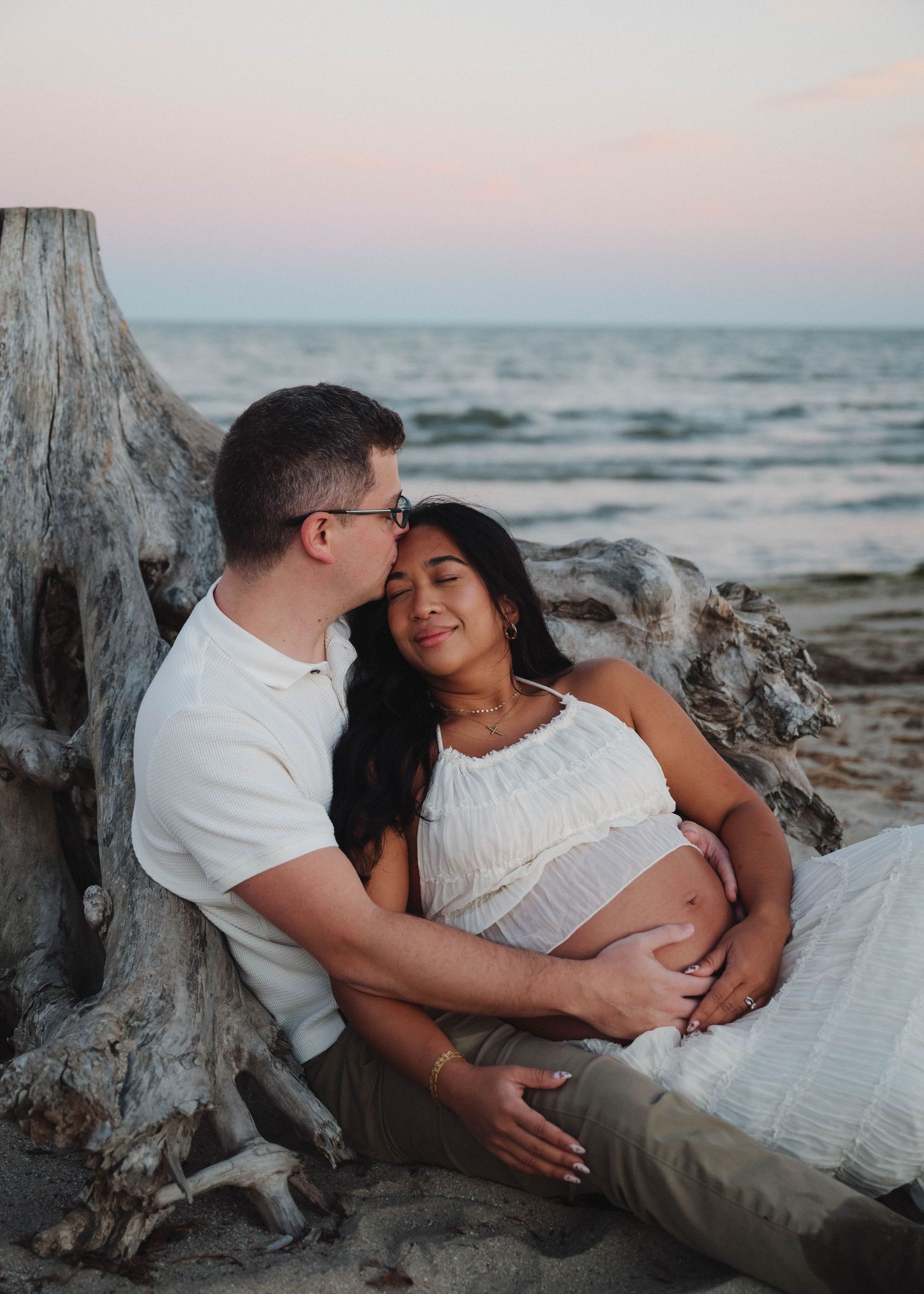 A couple shares an intimate moment on a beach at sunset, cuddling against a weathered piece of driftwood.