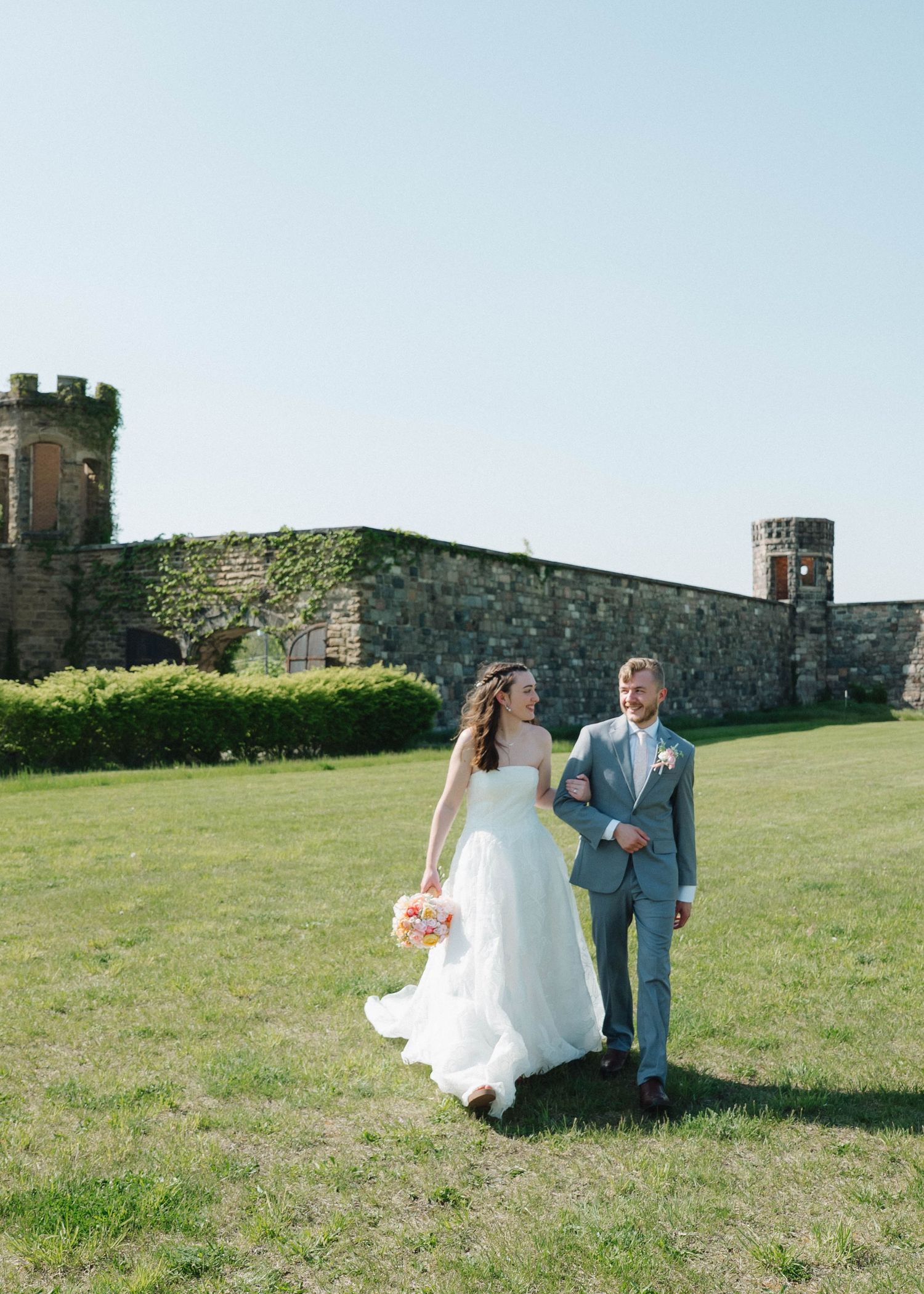 A couple in wedding attire walks across a grassy field near an old stone castle wall on a sunny day.