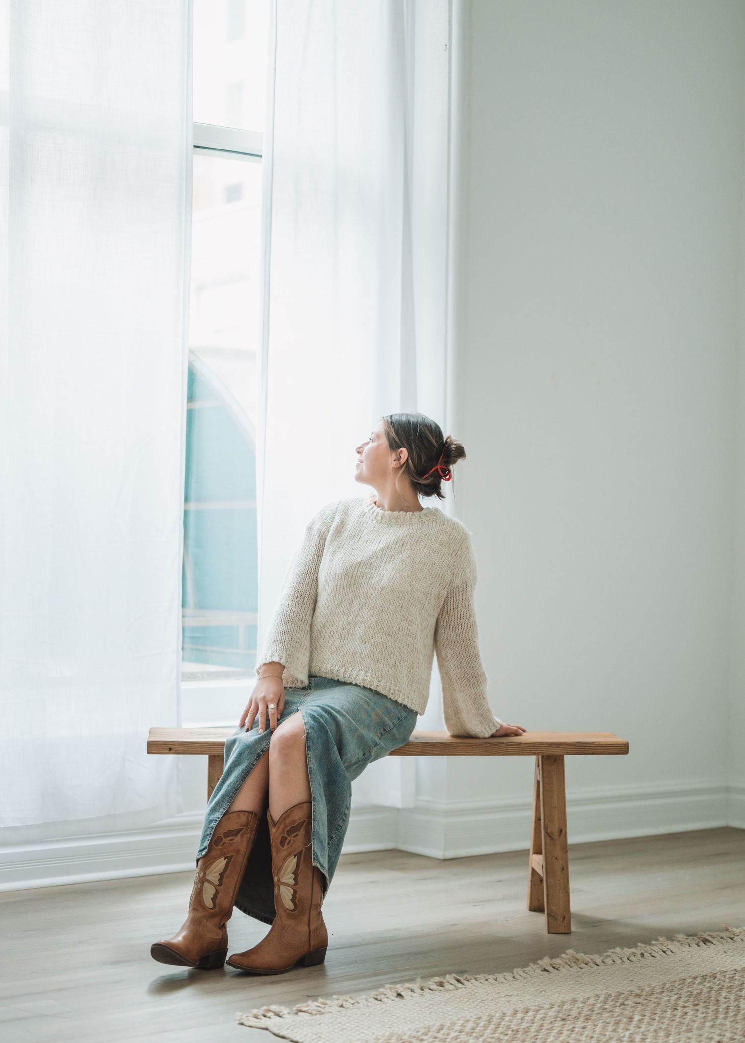 Someone in a cream sweater and brown boots sits thoughtfully on a wooden bench by a bright window with flowing curtains.