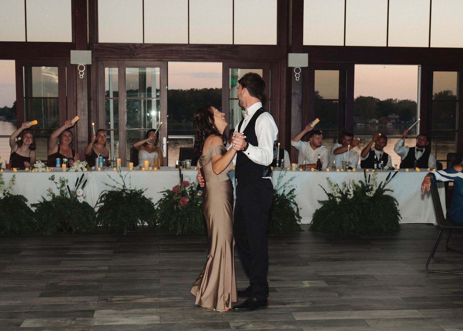 Wedding party members stand in formal attire on an outdoor deck at dusk.