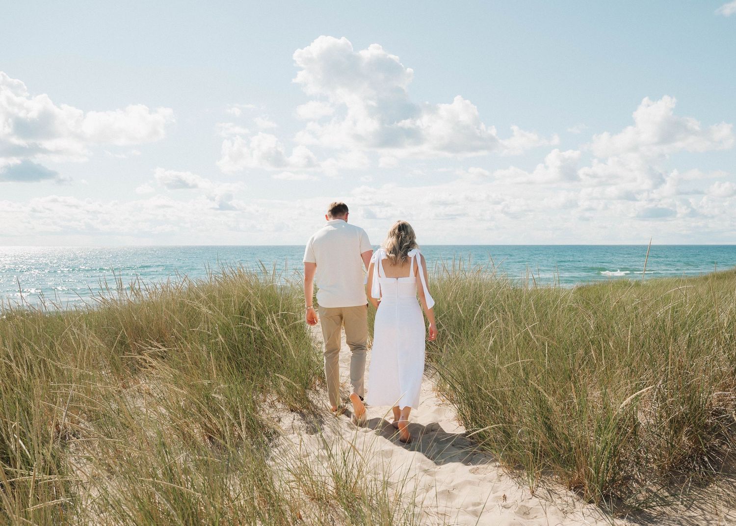 A couple walks along a sandy beach path through tall grass dunes with a bright blue ocean horizon in the distance.