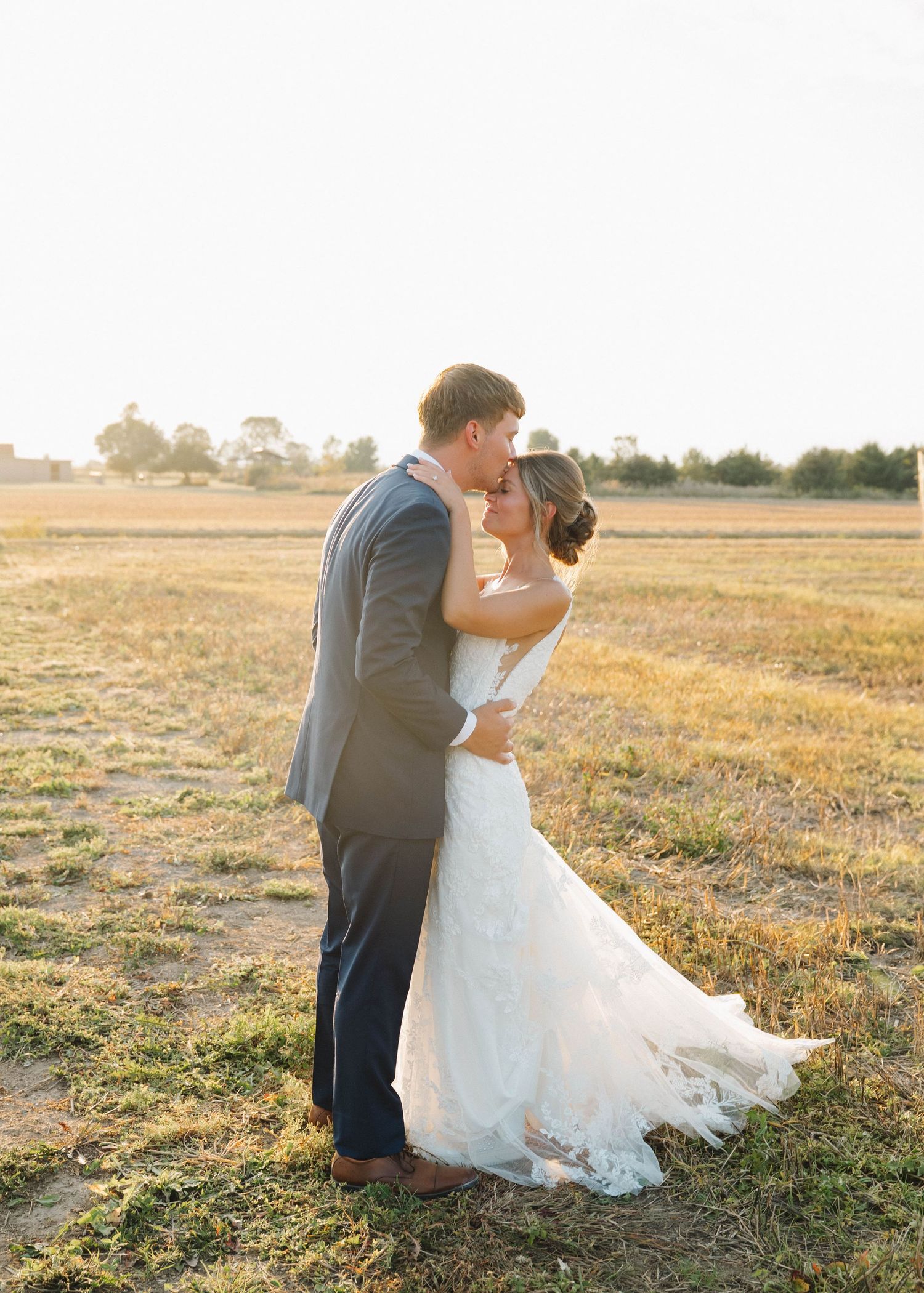 Romantic couple in wedding attire embraces in a golden sunlit field during sunset.