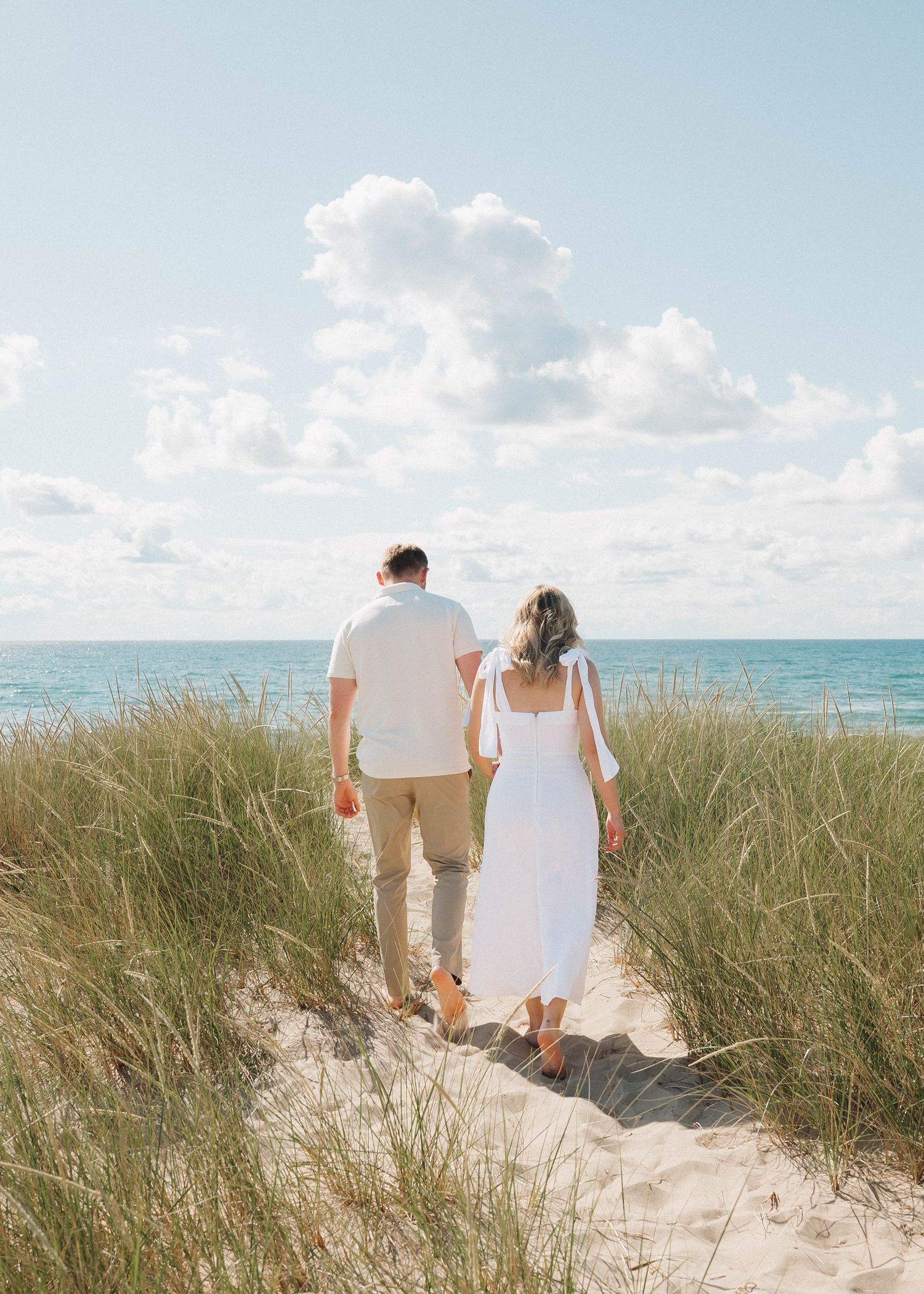 Couple walking hand in hand along a scenic beach path through tall sea grass on a sunny summer day.