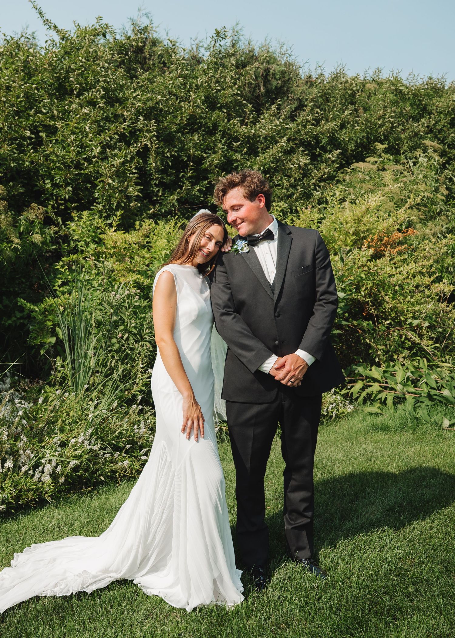 Bride in white gown and groom in black tuxedo pose together in lush garden on their wedding day.
