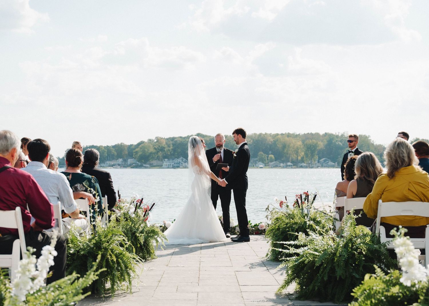 Panoramic view of an outdoor waterfront wedding ceremony with white floral arrangements and guests gathered by the shore.