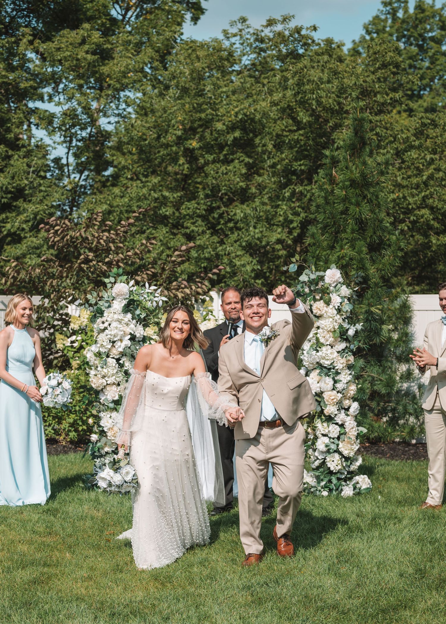 Couple exits outdoor wedding ceremony under floral arch with attendants in blue dresses.