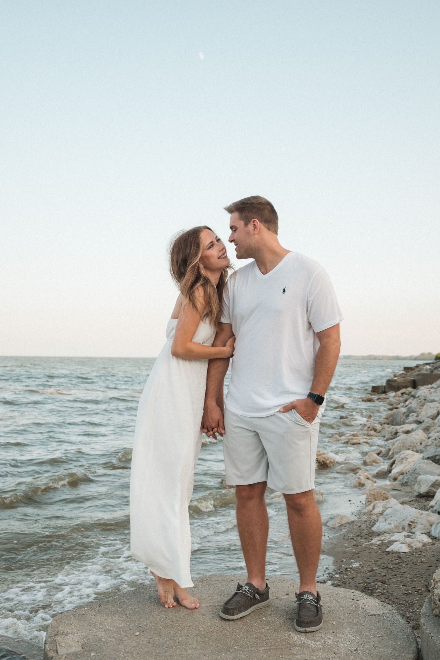 Romantic couple poses together on a rocky beach shoreline at sunset wearing white outfits.