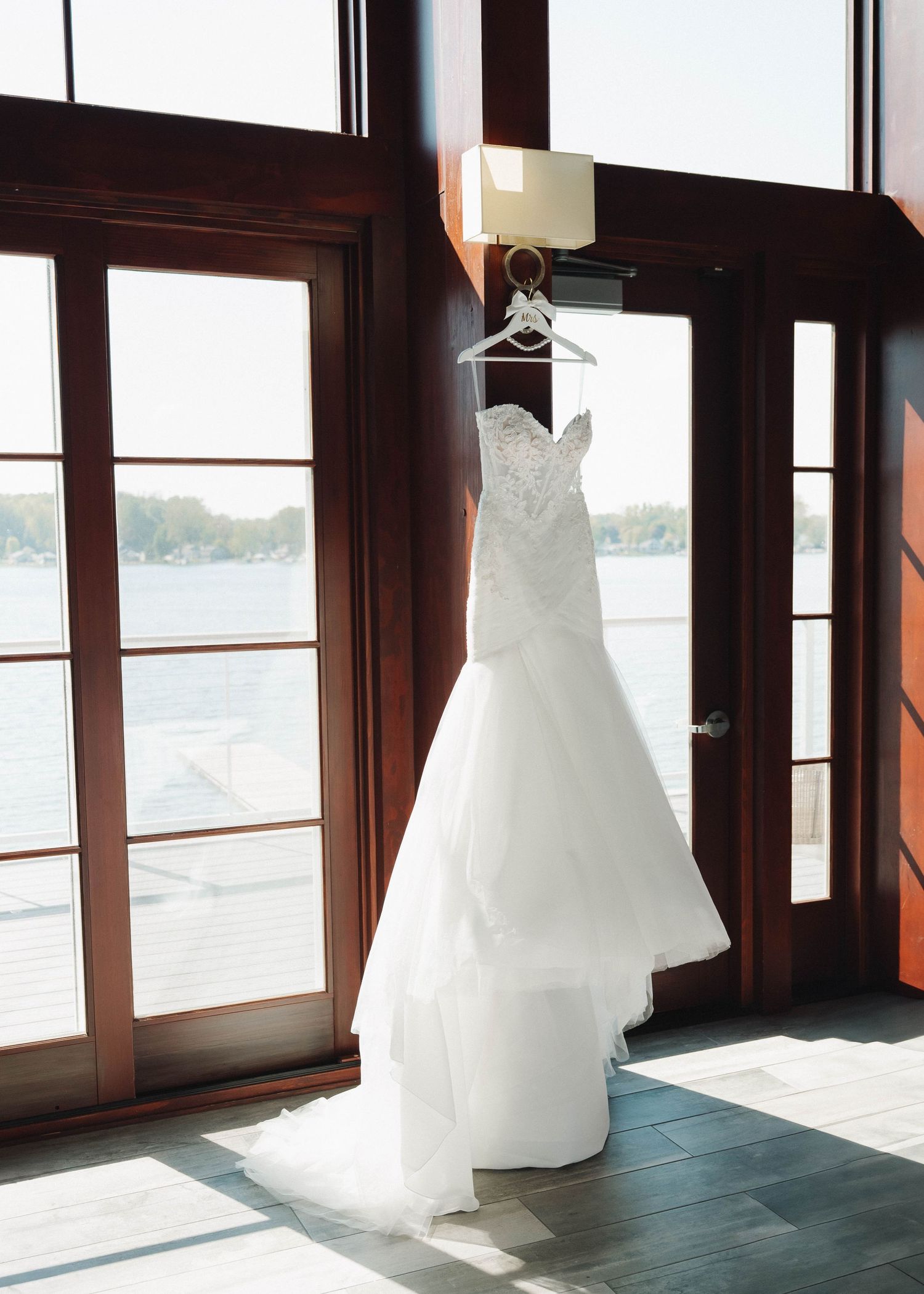 A white lace wedding dress hangs elegantly by a window with sunlight streaming in, displaying its mermaid silhouette.