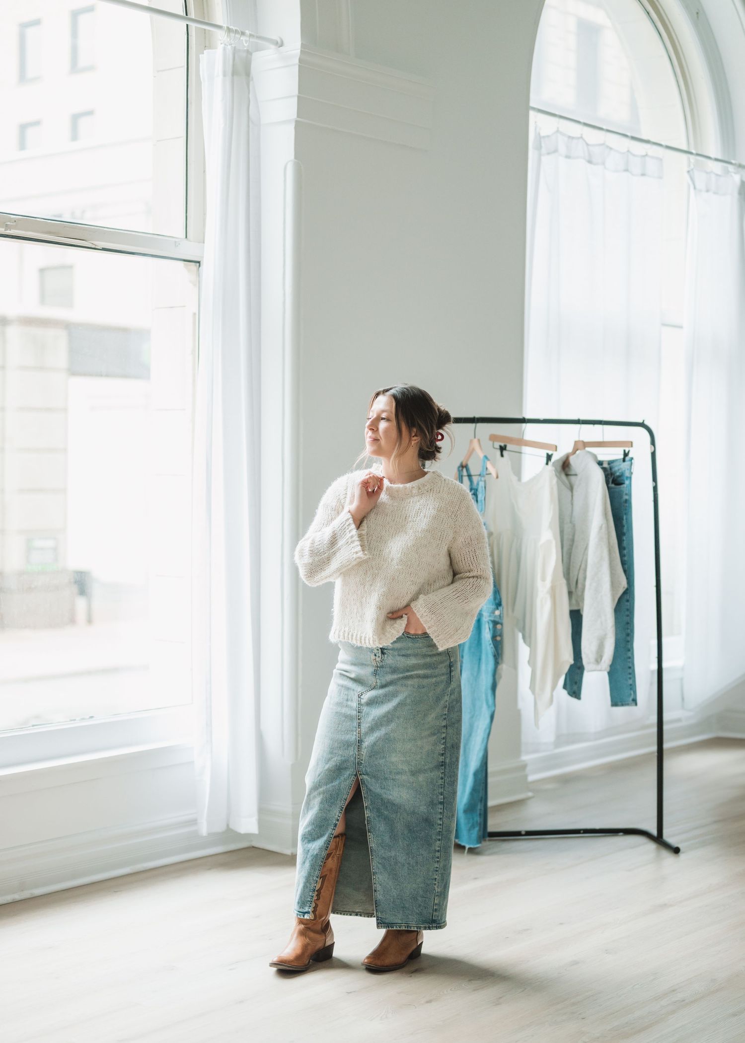 Person in denim and cream sweater posing casually in bright white studio with arched windows.