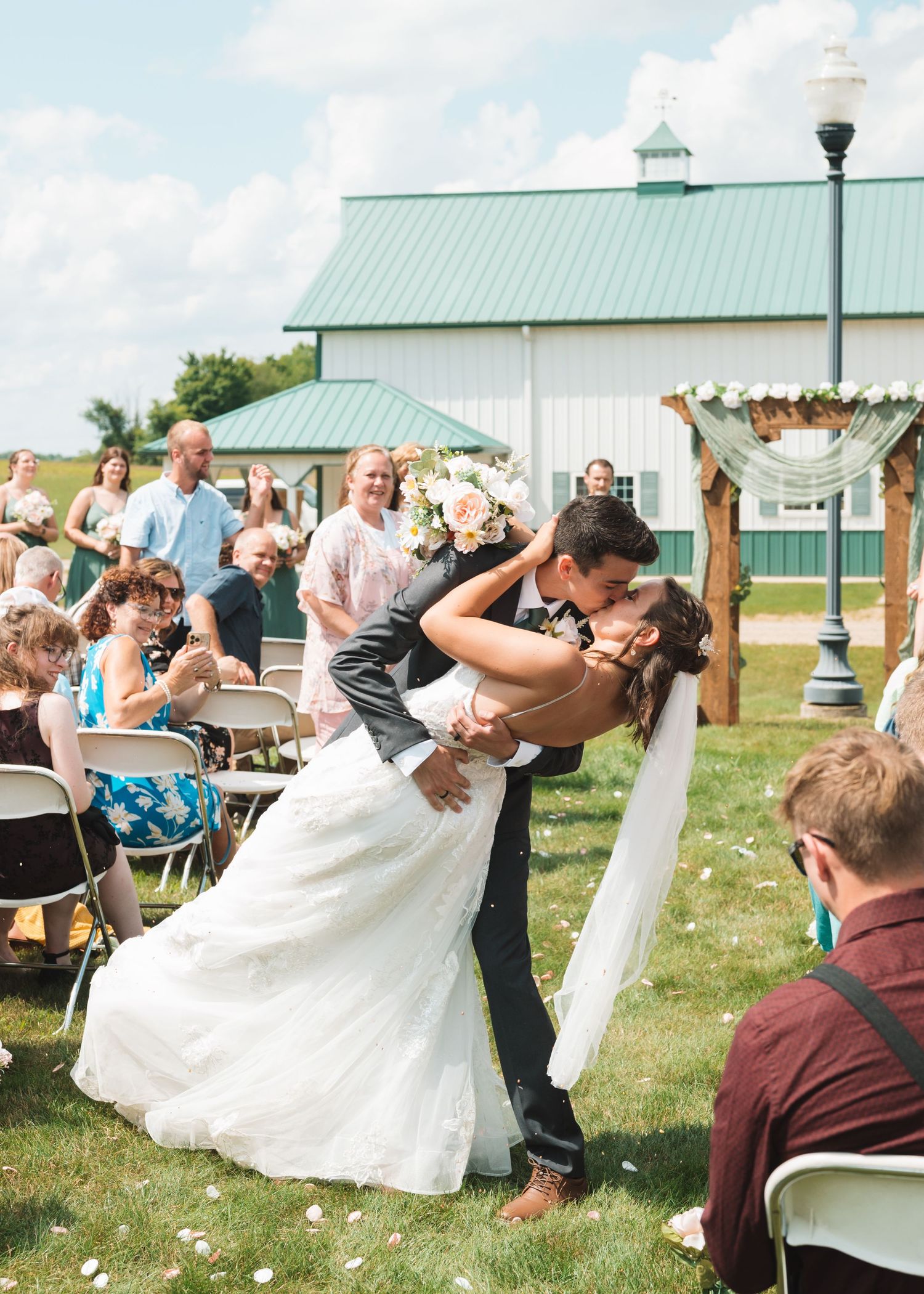A classic wedding dip kiss performed in front of a white barn with guests looking on.