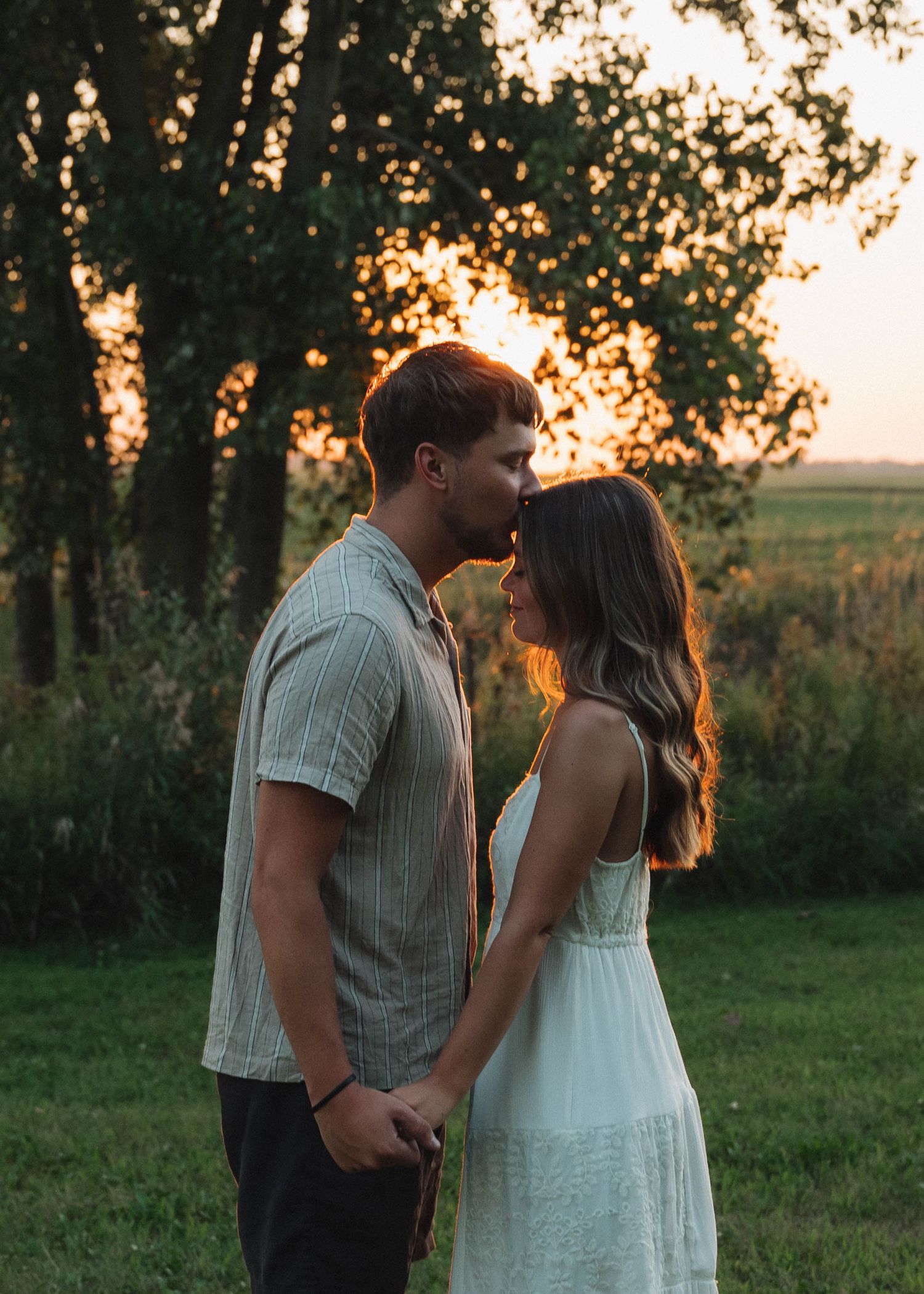 A couple shares a tender moment at sunset in a grassy field with trees silhouetted in the background.