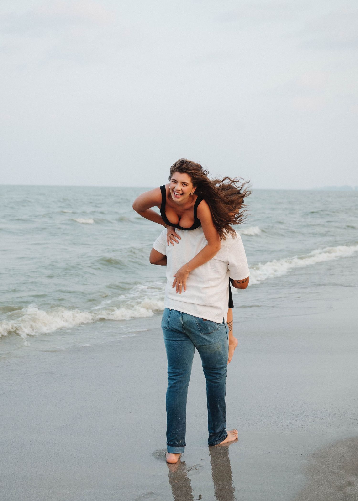 A person in jeans and white shirt walks joyfully along a misty beach shoreline with waves in the background.