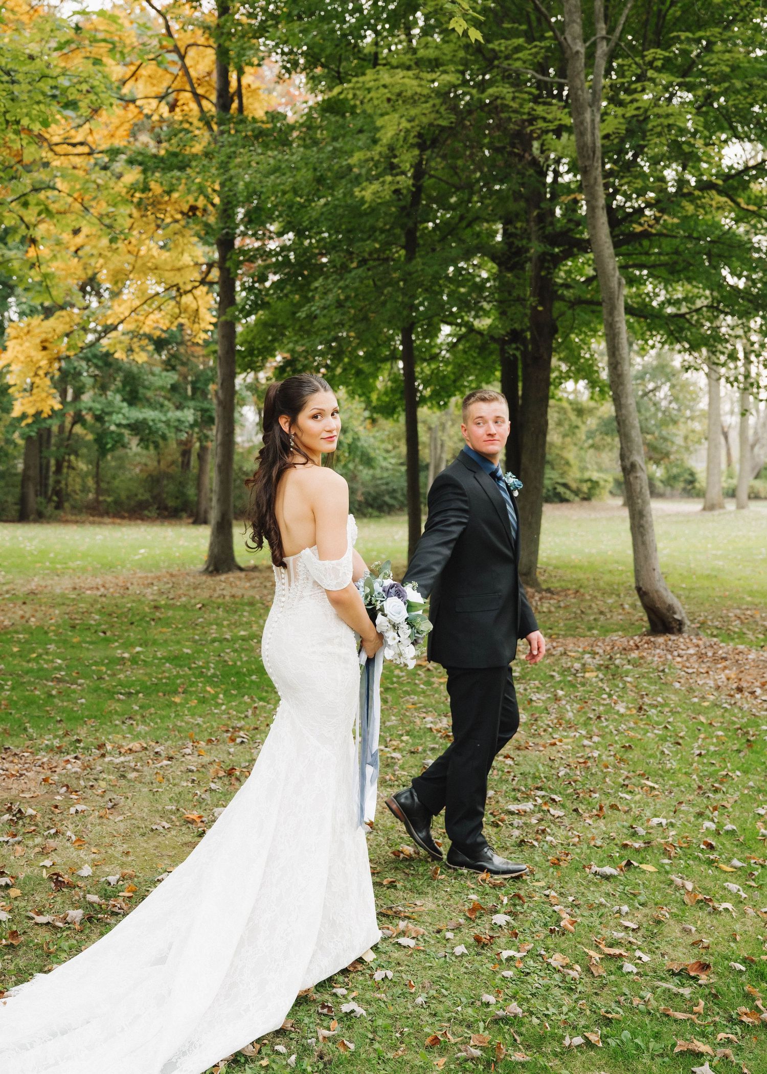 A couple in wedding attire walks together through a scenic autumn park with fallen leaves on the ground.