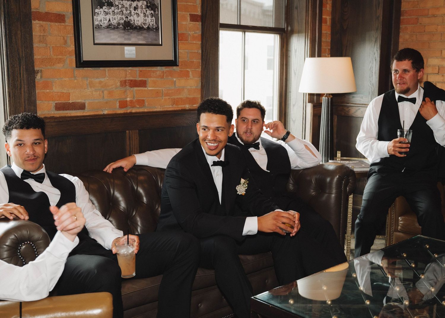 Groomsmen in black suits gather at a rustic brick-walled venue for wedding celebration drinks.