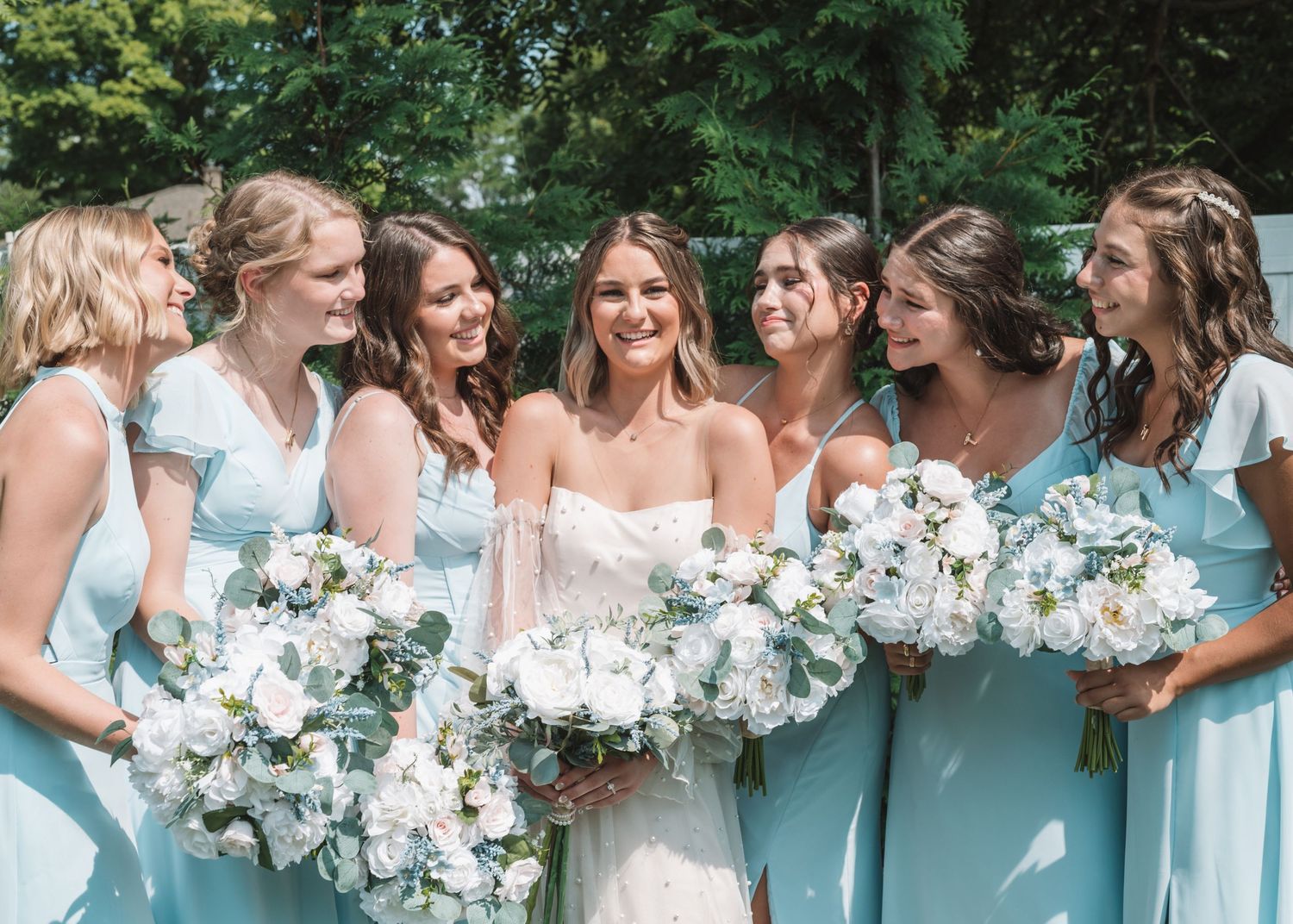 Bridesmaids in light blue dresses hold white floral bouquets while sharing joyful moment.