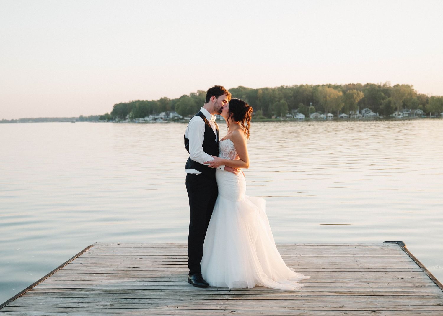 A romantic wedding couple embraces on a wooden dock overlooking a serene lake at sunset.