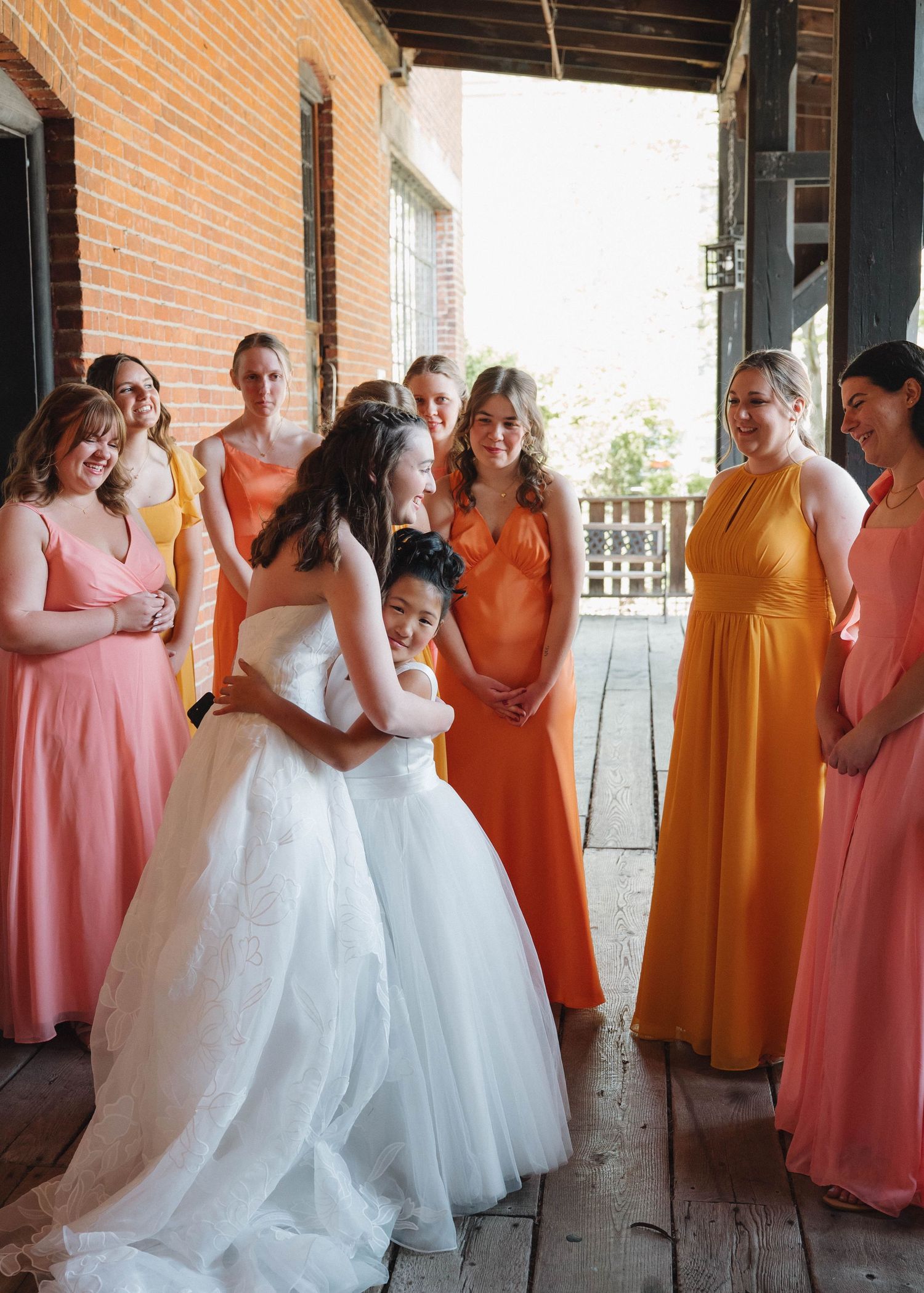 Wedding party in orange and coral bridesmaid dresses lined up along a brick wall for candid photos.
