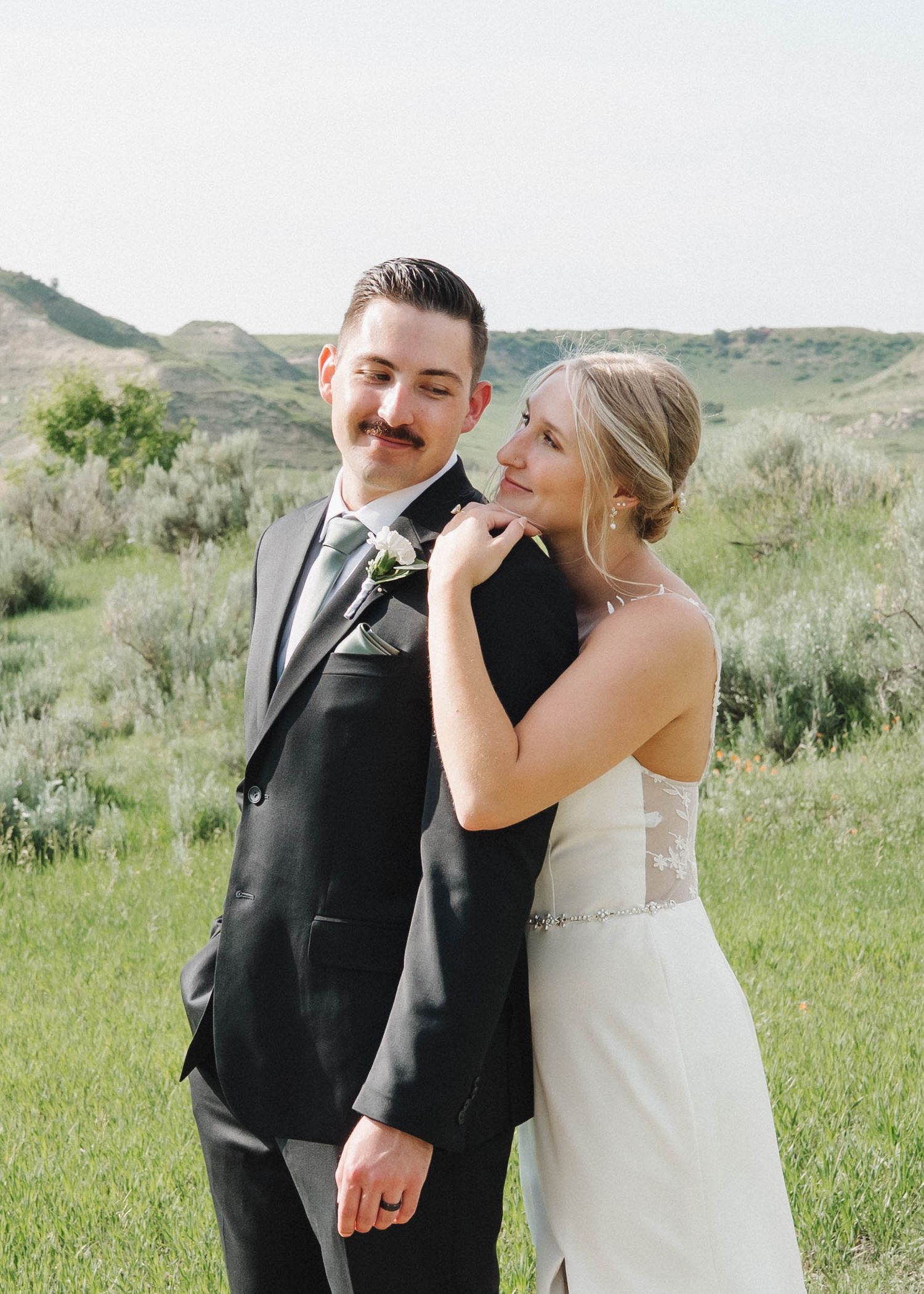 A bride and groom share a playful moment together in a green meadow with rolling hills in the background.