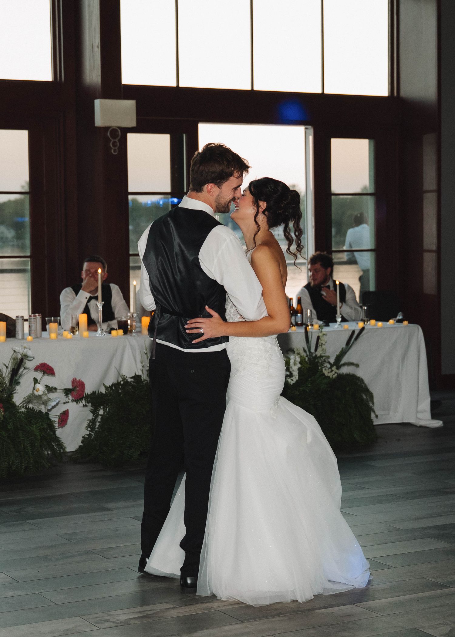 A couple shares their first dance together at their wedding reception with floral decor and tables in background.