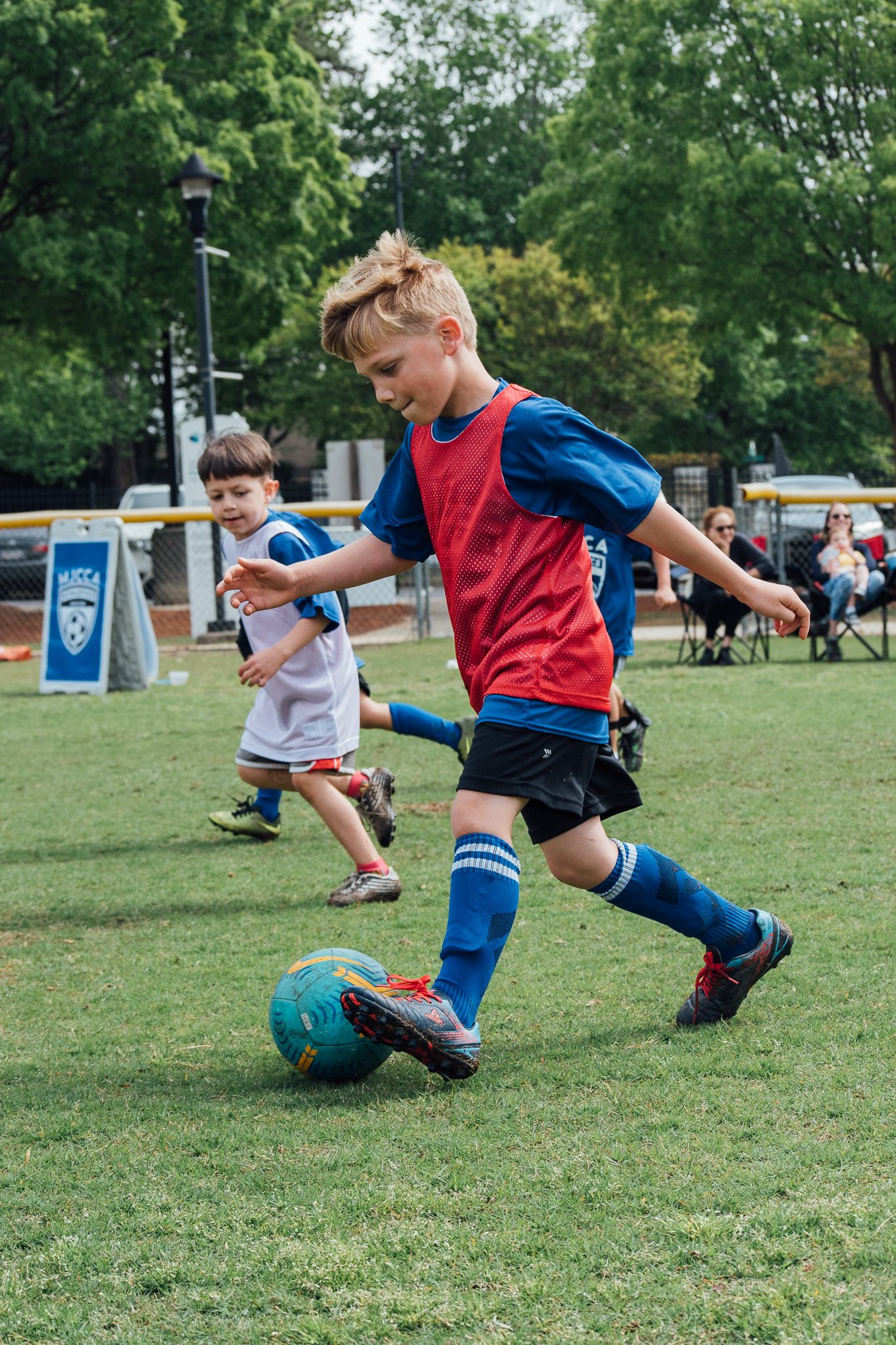 Young boy in red vest dribbling soccer ball during youth league match on green grass field.