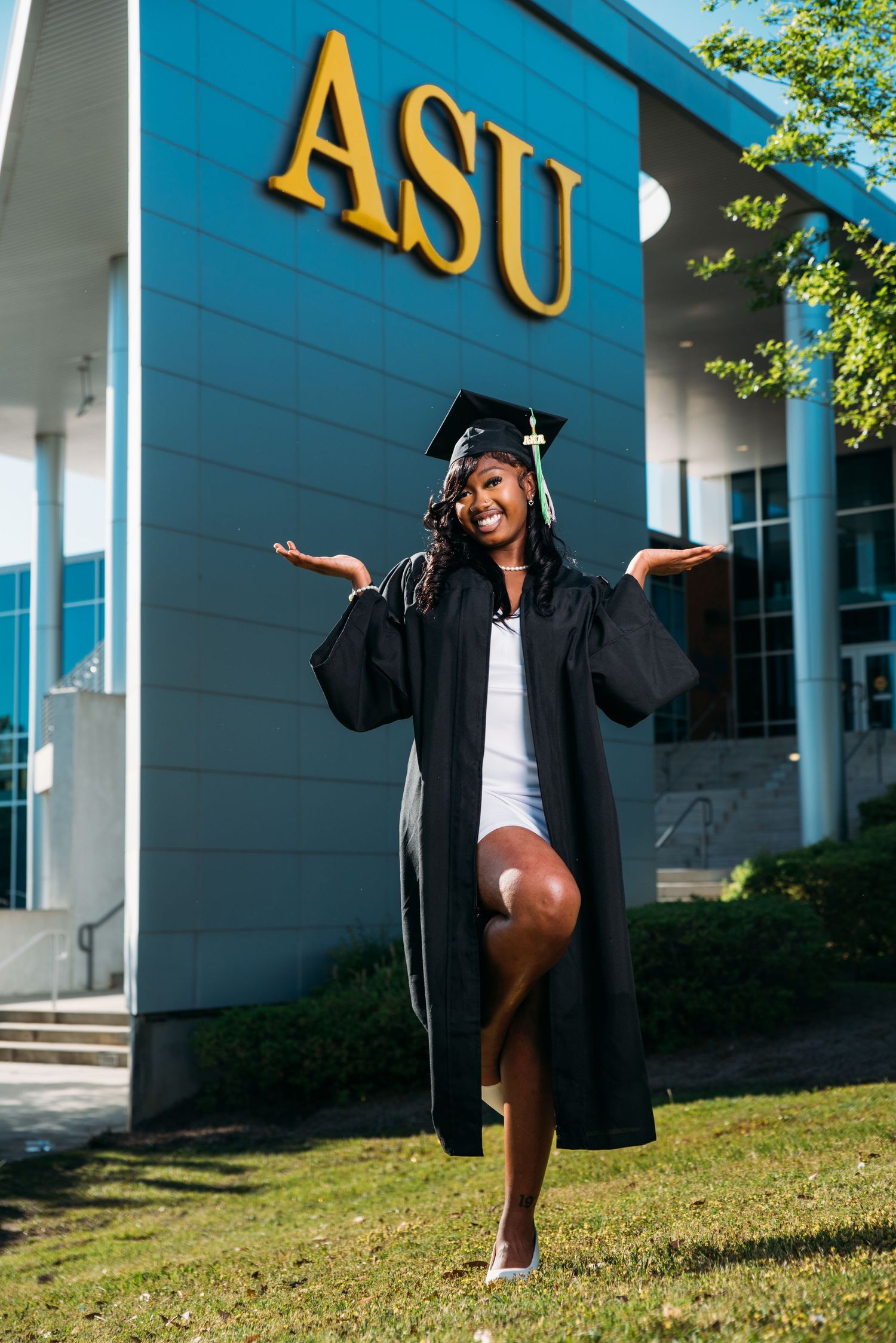 Graduate in cap and gown poses joyfully in front of blue building with yellow signage.