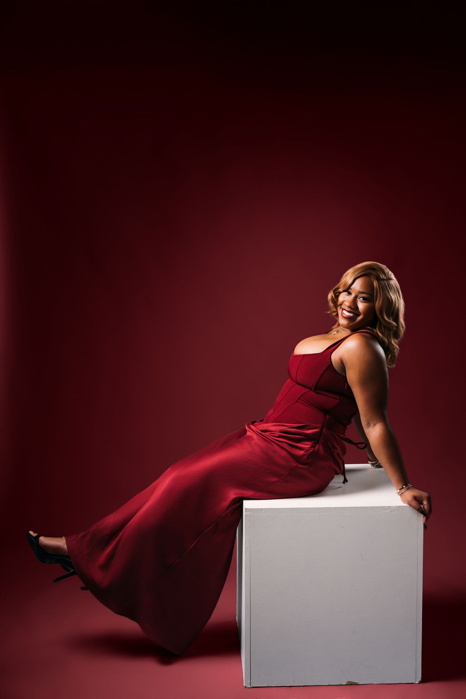 A smiling woman in a red gown poses on a white cube against a deep red background.