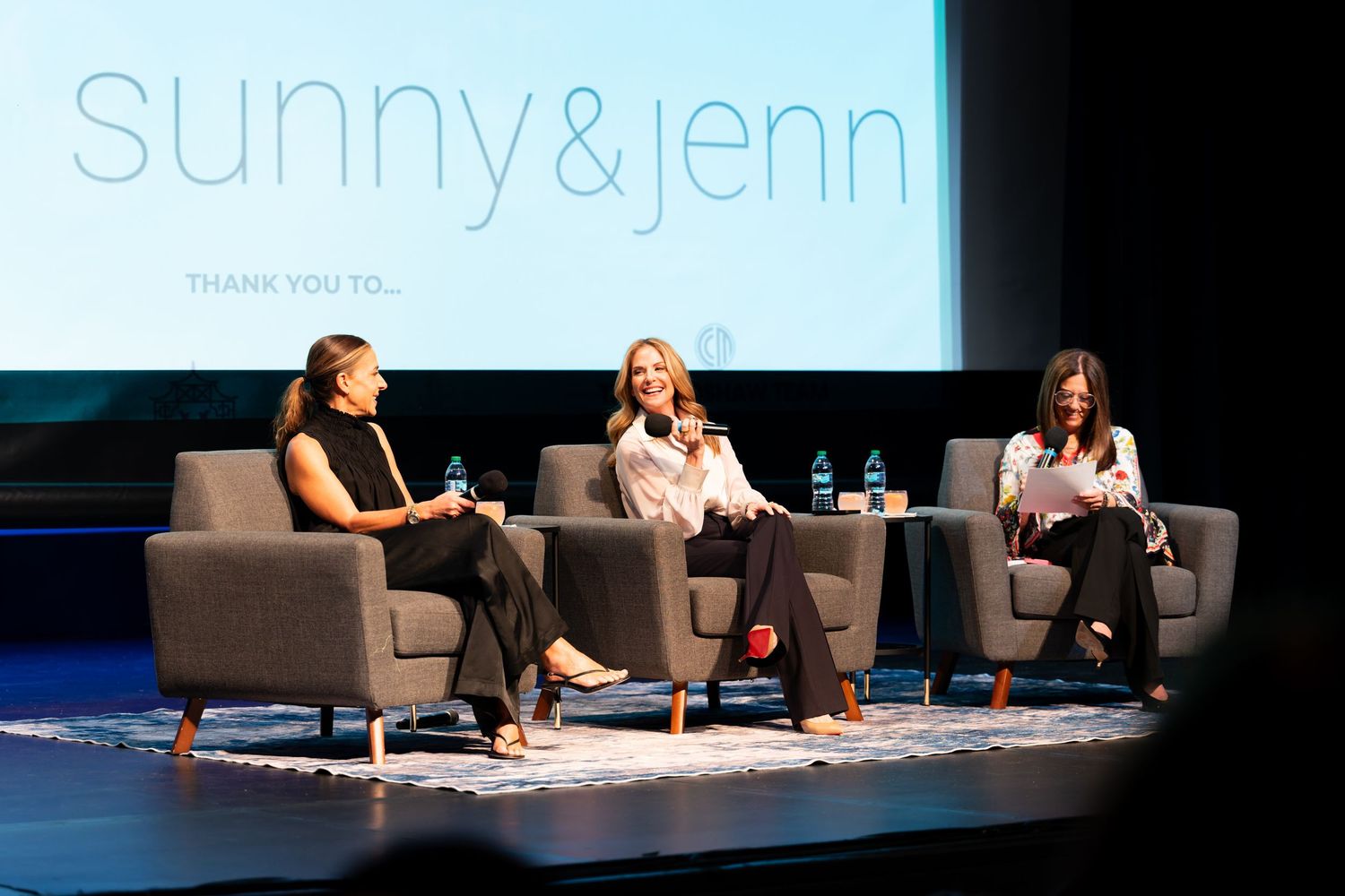 Three women sit in gray chairs on stage at a Sunny & Jenn event, engaged in a panel discussion.
