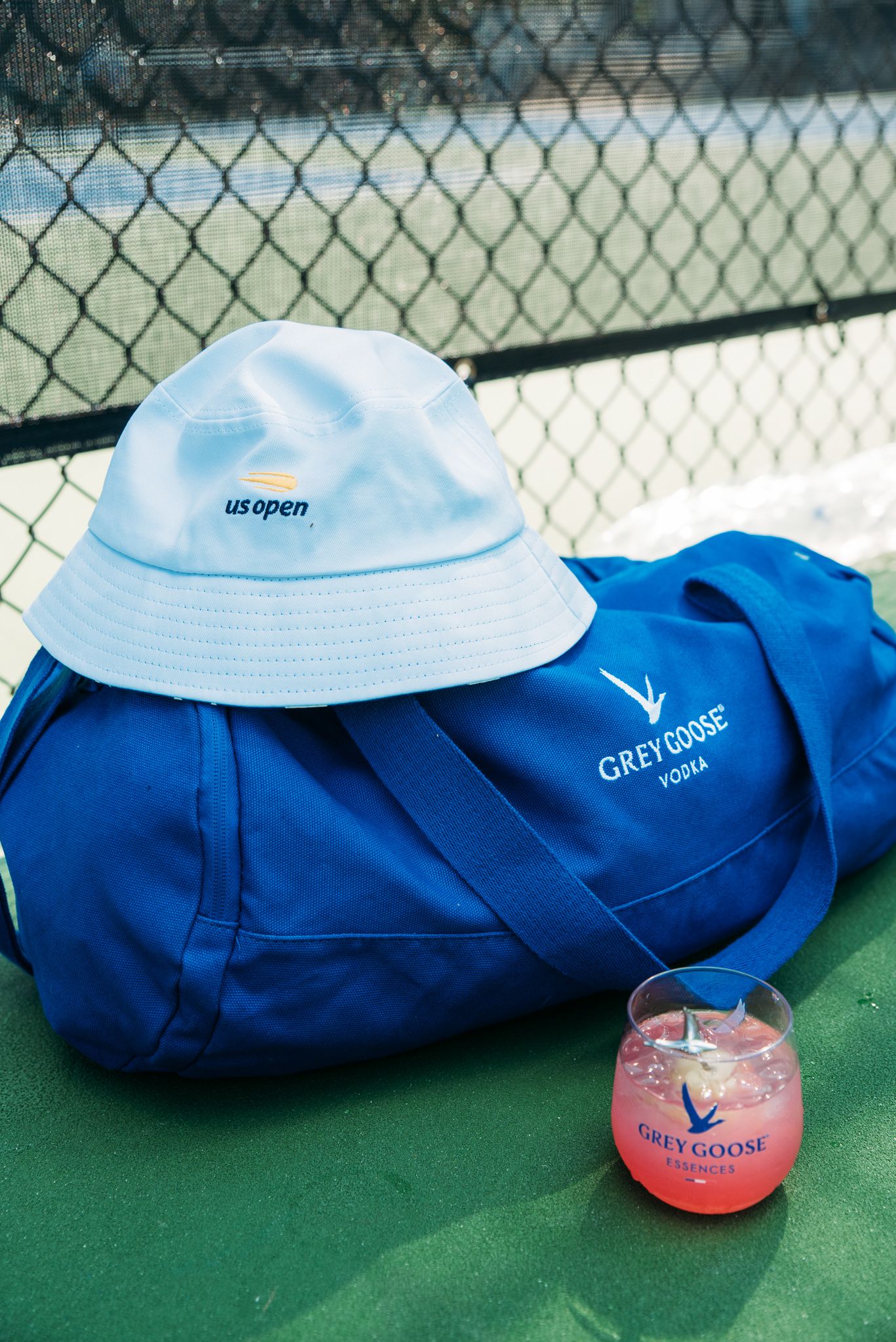 Blue duffle bag and light blue bucket hat with pink drink on tennis court.