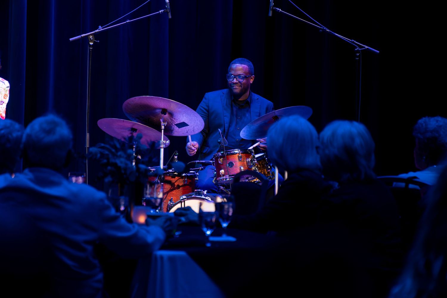 Drummer performs on stage with atmospheric blue lighting and silhouetted audience.