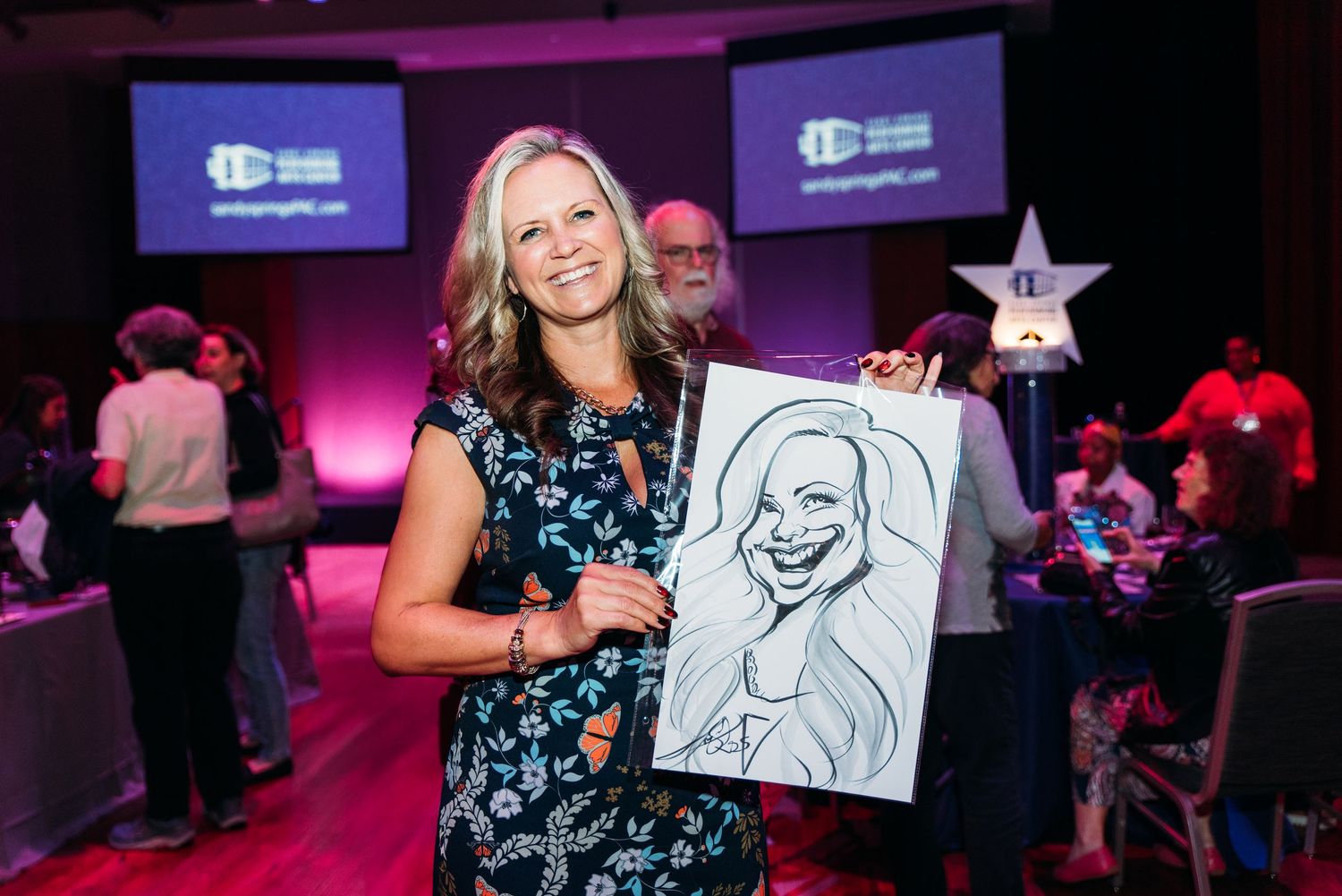 Event attendees displaying caricature drawings at a dimly lit venue.