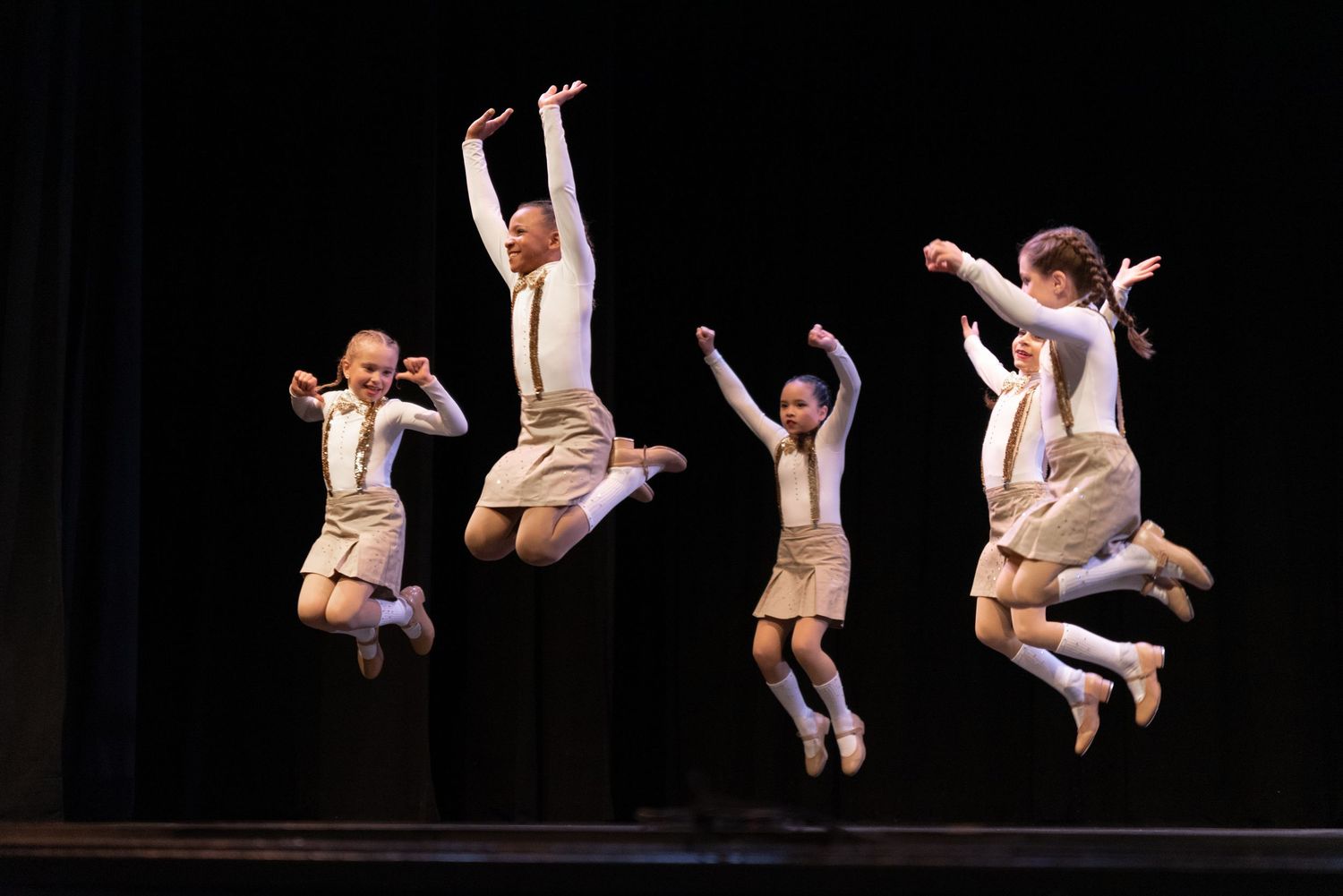 Ballet dancers in white dresses leaping gracefully across a dark stage during a performance.
