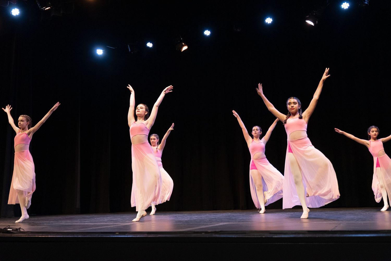 Group of dancers in flowing white dresses performing synchronized movements under stage lighting.