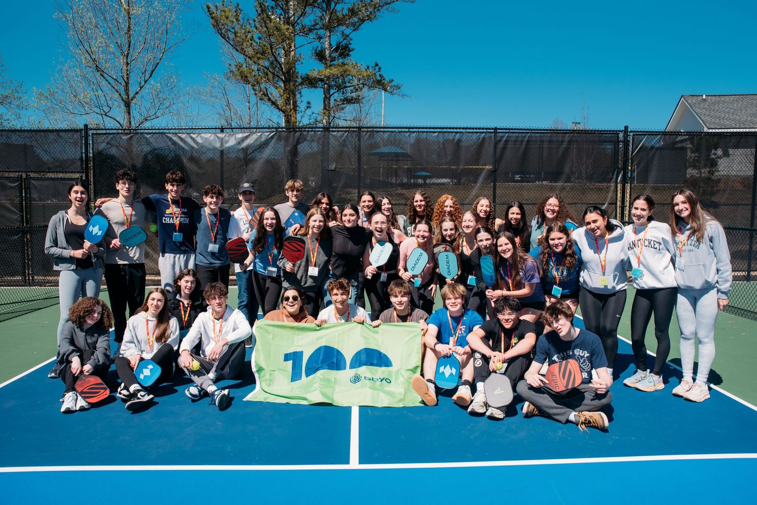 Large group gathered outdoors on a sunny day for a group photo on tennis courts.