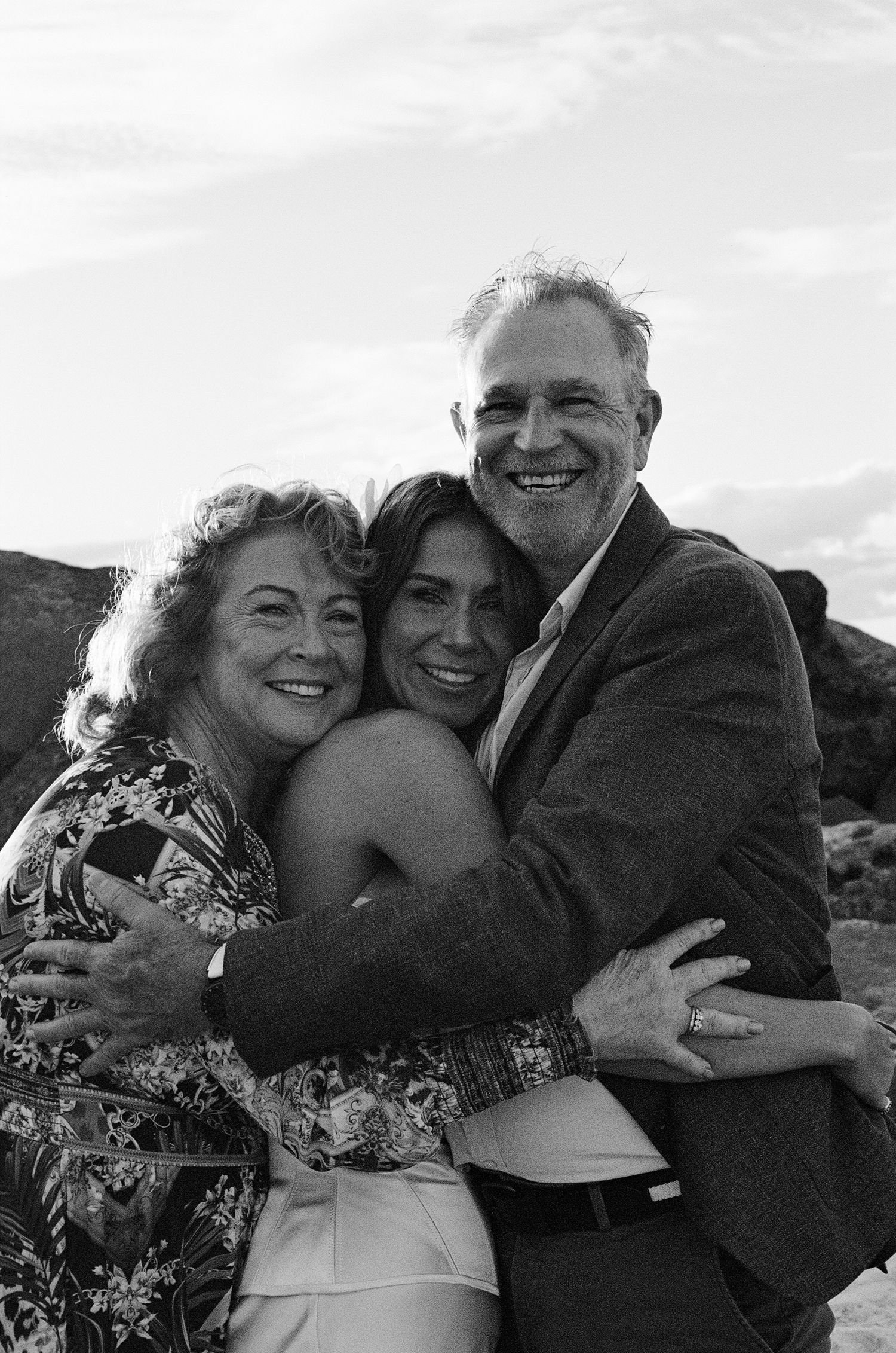 A black and white outdoor portrait of three people embracing and smiling together against a mountain backdrop.