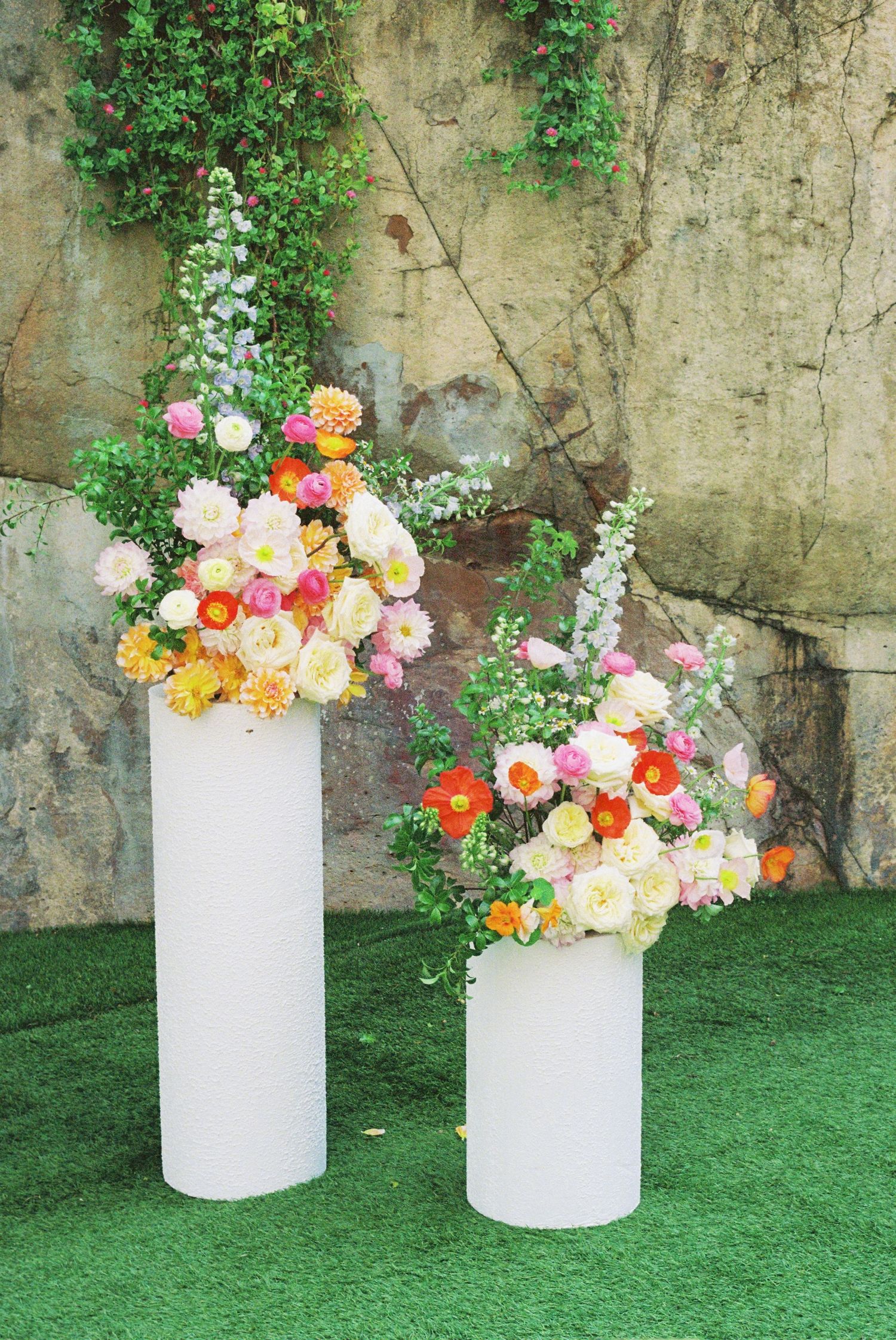 Two elegant white pedestals adorned with colorful summer flowers against a textured stone wall.