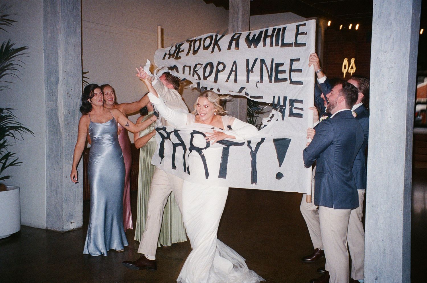 Wedding guests dance energetically under a handmade banner at a lively reception celebration.