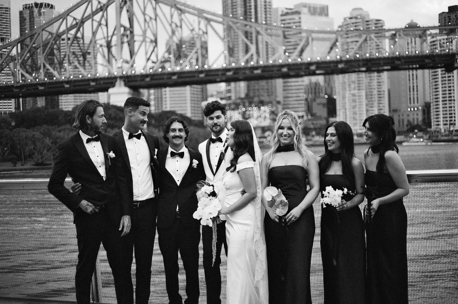Wedding party wearing formal attire poses along a scenic waterfront with a steel bridge in the background.