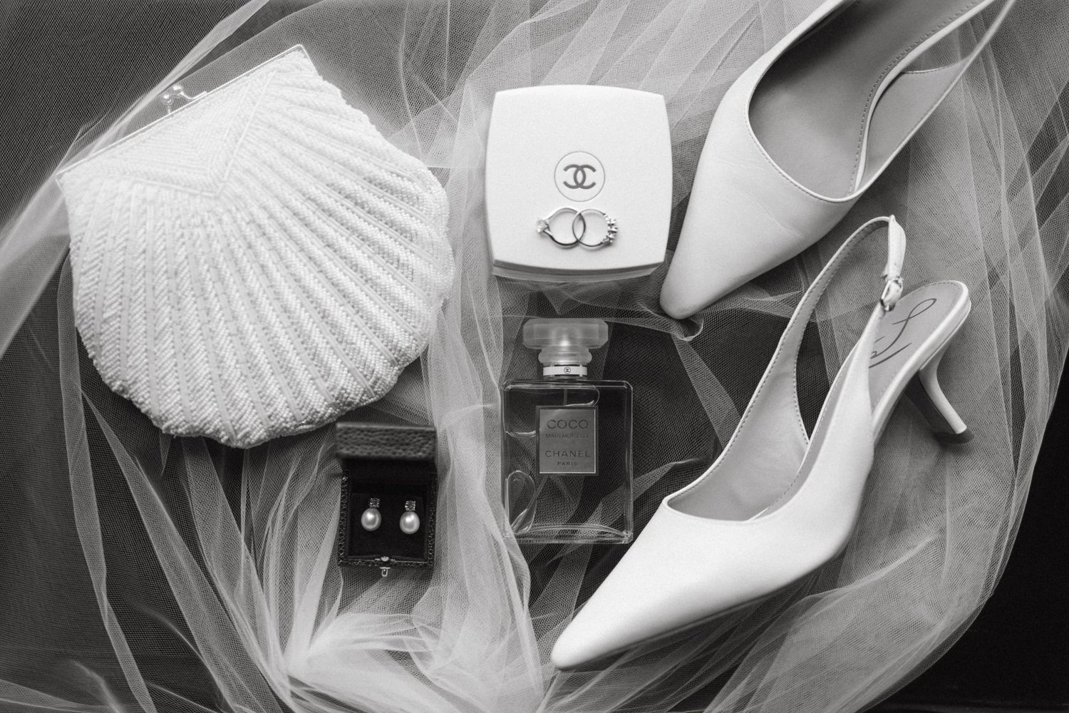 A wedding themed flat lay featuring white heels, seashells, pearls, and jewelry arranged on white tulle fabric.