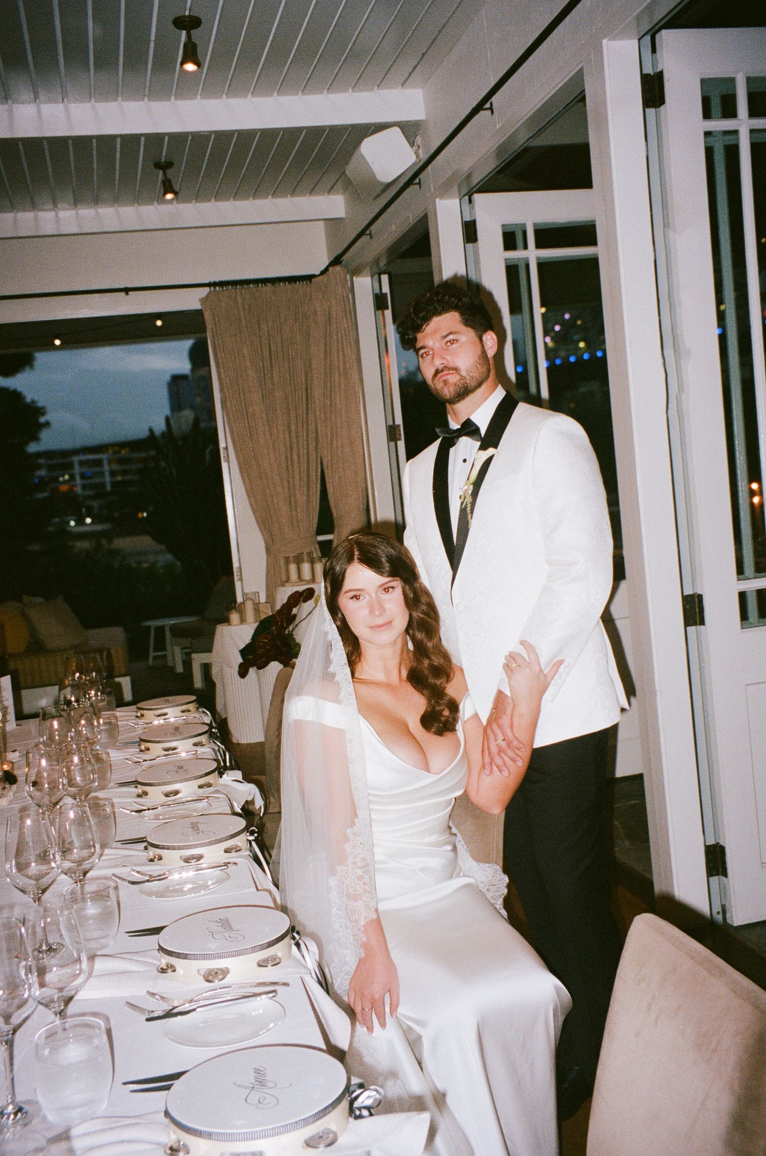 A couple poses together at an elegant wedding reception dinner table set with wine glasses.
