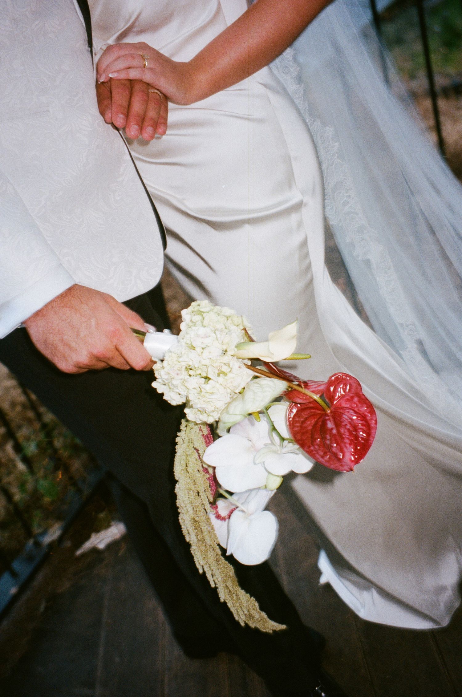 A close-up of a wedding bouquet with white flowers and red anthurium blooms against a white dress.
