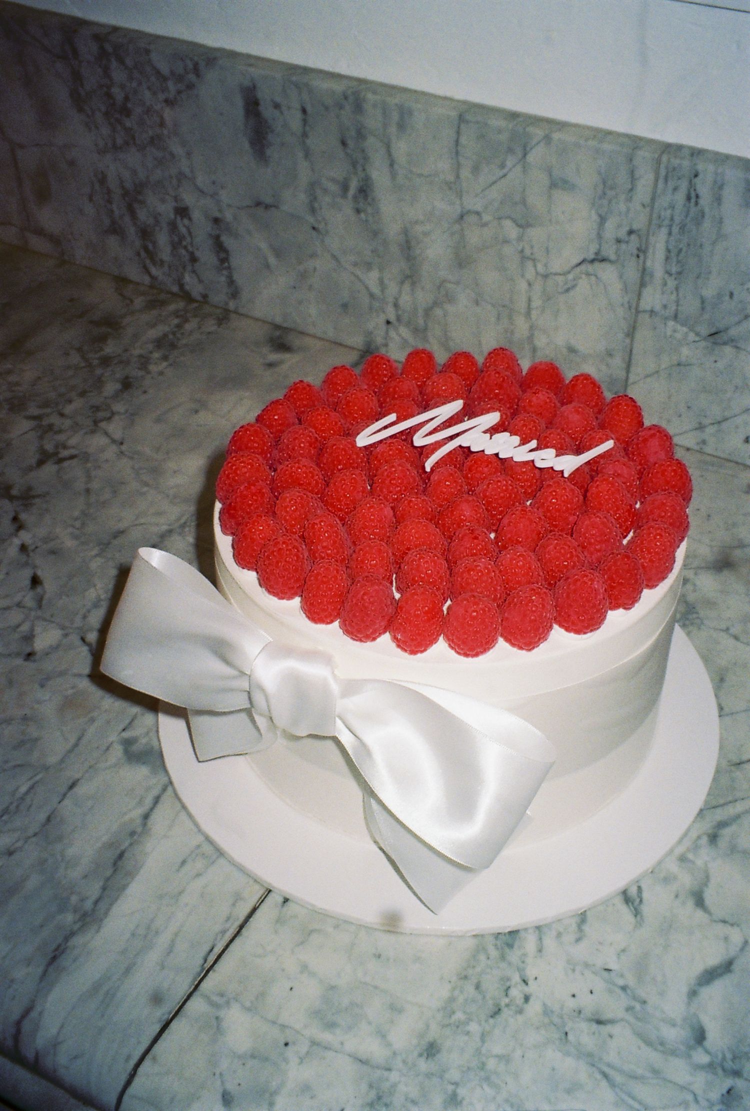 A white round cake decorated with red raspberries and a white bow ribbon around the base.