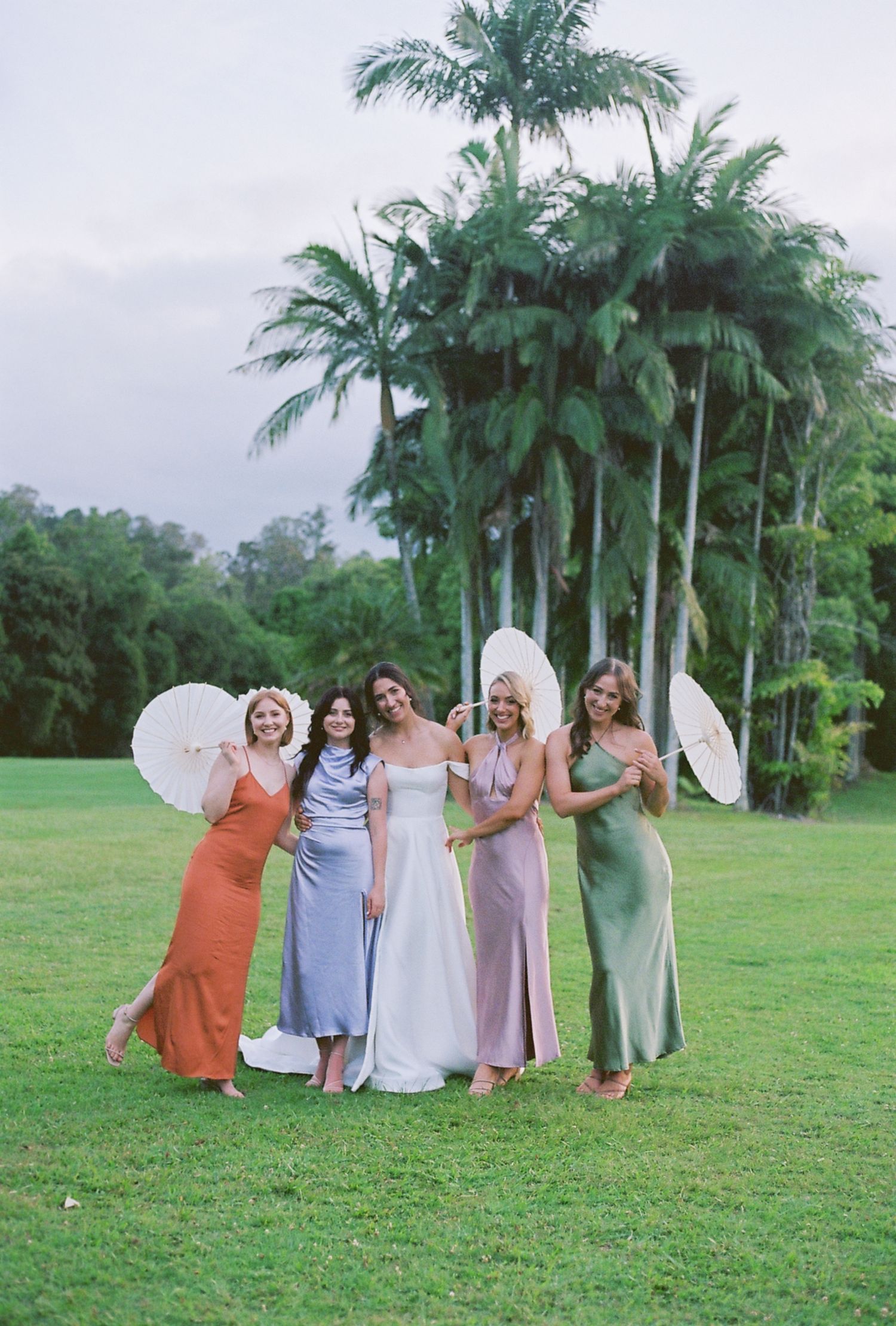 A bridal party poses with parasols against a backdrop of towering palm trees.