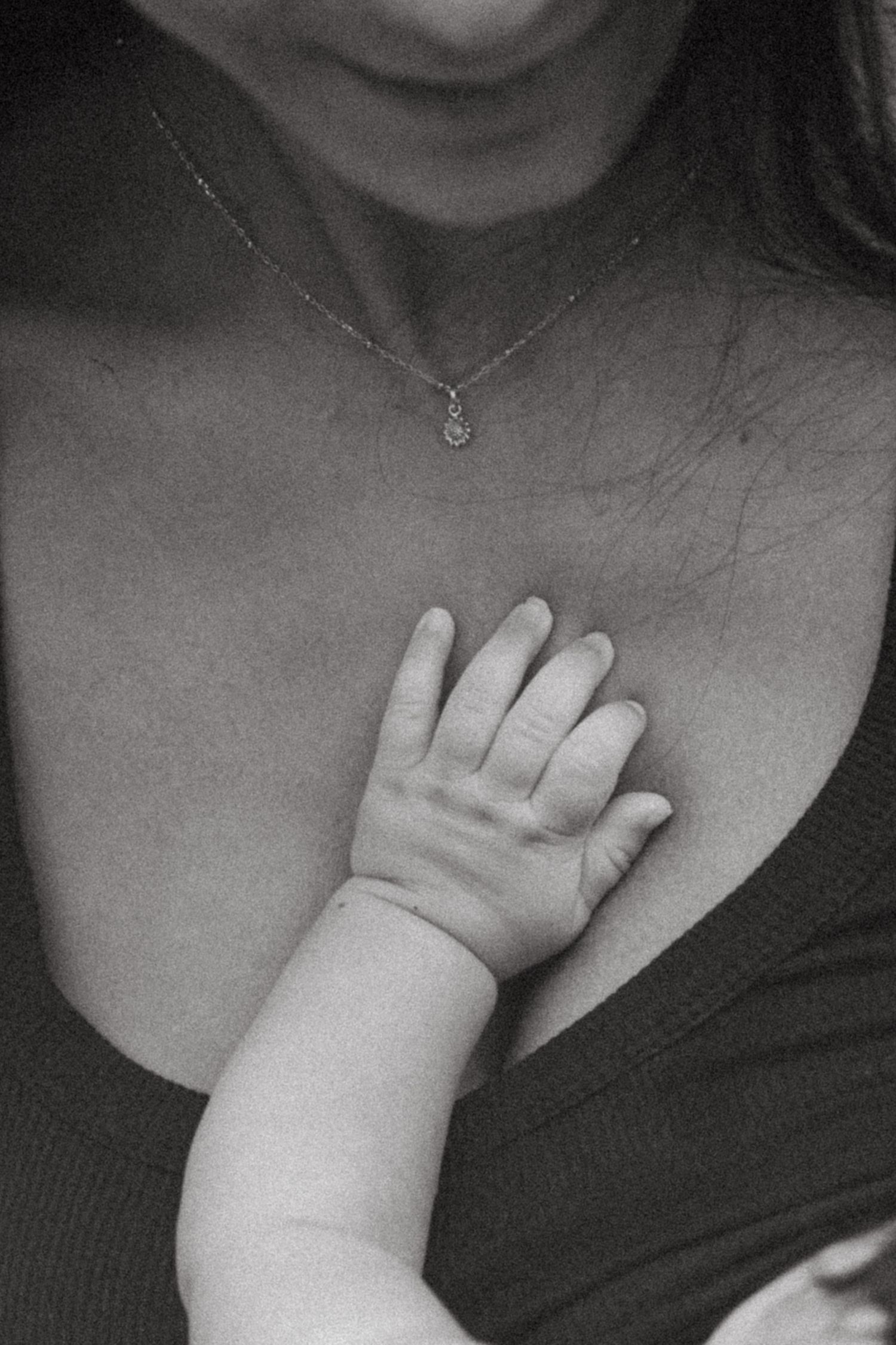 Close-up black and white image of baby's hand resting on parent's chest with delicate necklace.